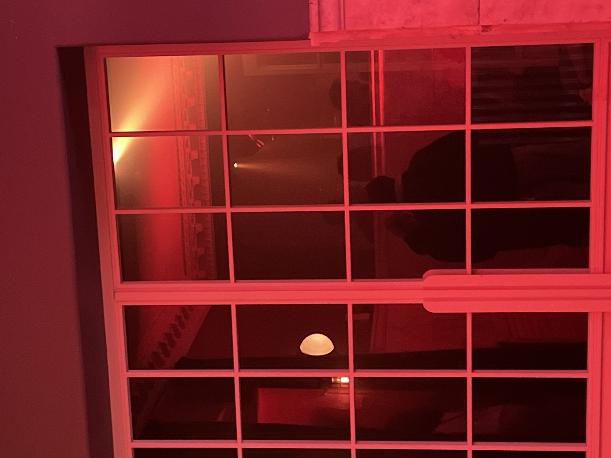 A window with multiple panes reflects a red-lit room, partially obscuring the view outside. Reflected within the window are interior details, including wall decor and furniture. The lighting is predominantly red, suggesting an artificial or colored light source. Outside the window, the reflection suggests another building with blinds. The location is indiscernible from the image.