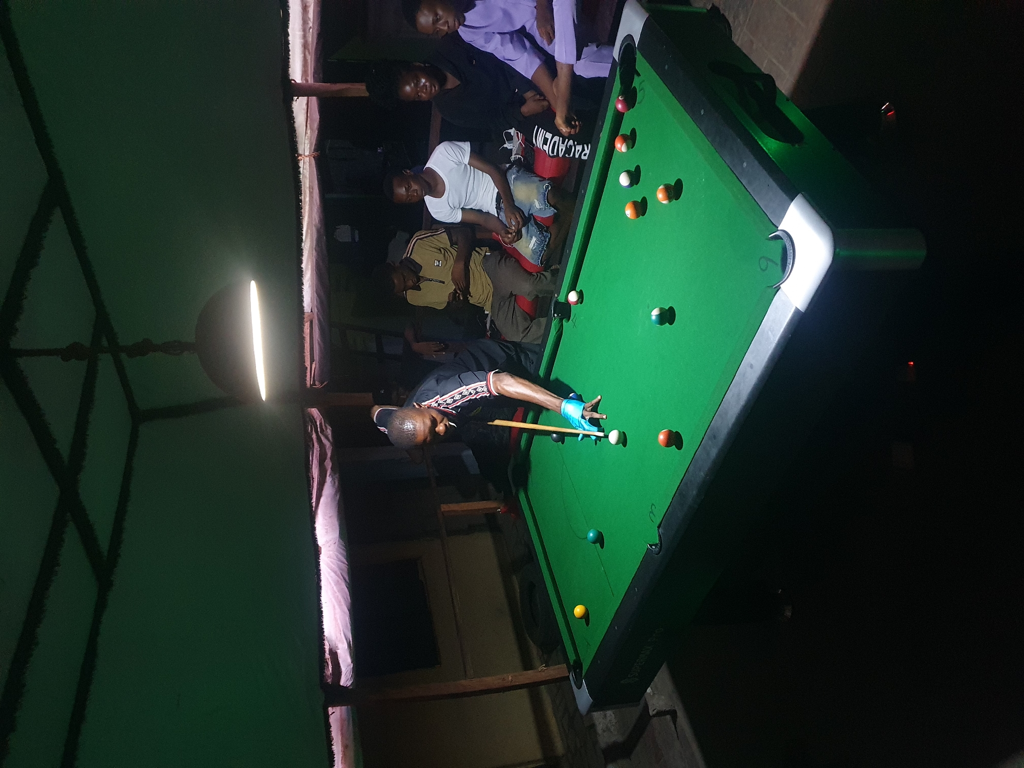 An indoor scene in Lokoja, Nigeria, captures a group of men engaged in and observing a game of pool under dim, artificial lighting, suggesting evening or night. In the foreground, a man wearing a black and patterned short-sleeved shirt and a light blue glove on his left hand leans over a green-felt pool table, lining up a shot with a cue stick. The cue ball and several other colored and striped pool balls are scattered across the table.
In the background, several other men are seated on a bench, observing the game. One man wears a white t-shirt and blue jeans, another a brown collared shirt, and two others are partially visible, one in a light purple top. The atmosphere appears casual.
A prominent, elongated light fixture hangs from the ceiling, brightly illuminating the playing area of the pool table. Visible text includes "RACADEMY" on a red bucket placed near the bench, and partially, "SUPERSNA PRO" on the side rail of the pool table. The setting suggests a local pool hall or a recreational area.