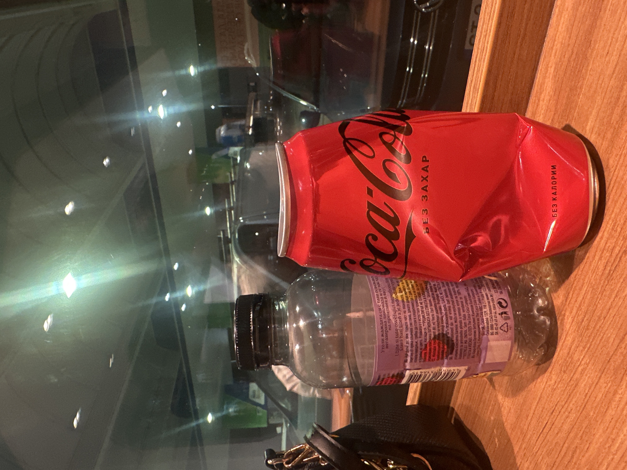 The image depicts a Coca-Cola Zero can and a transparent plastic bottle on a wooden surface near a window. The can is red with black lettering and appears slightly crushed. The bottle has a black cap and a purple label. The background shows reflections in the glass, including a car. The text on the Coca-Cola can is written in Cyrillic. Part of the bottle label text references a location in Bulgaria. Therefore, it can be inferred this image was taken somewhere in Bulgaria, but not specifically Stara Zagora.