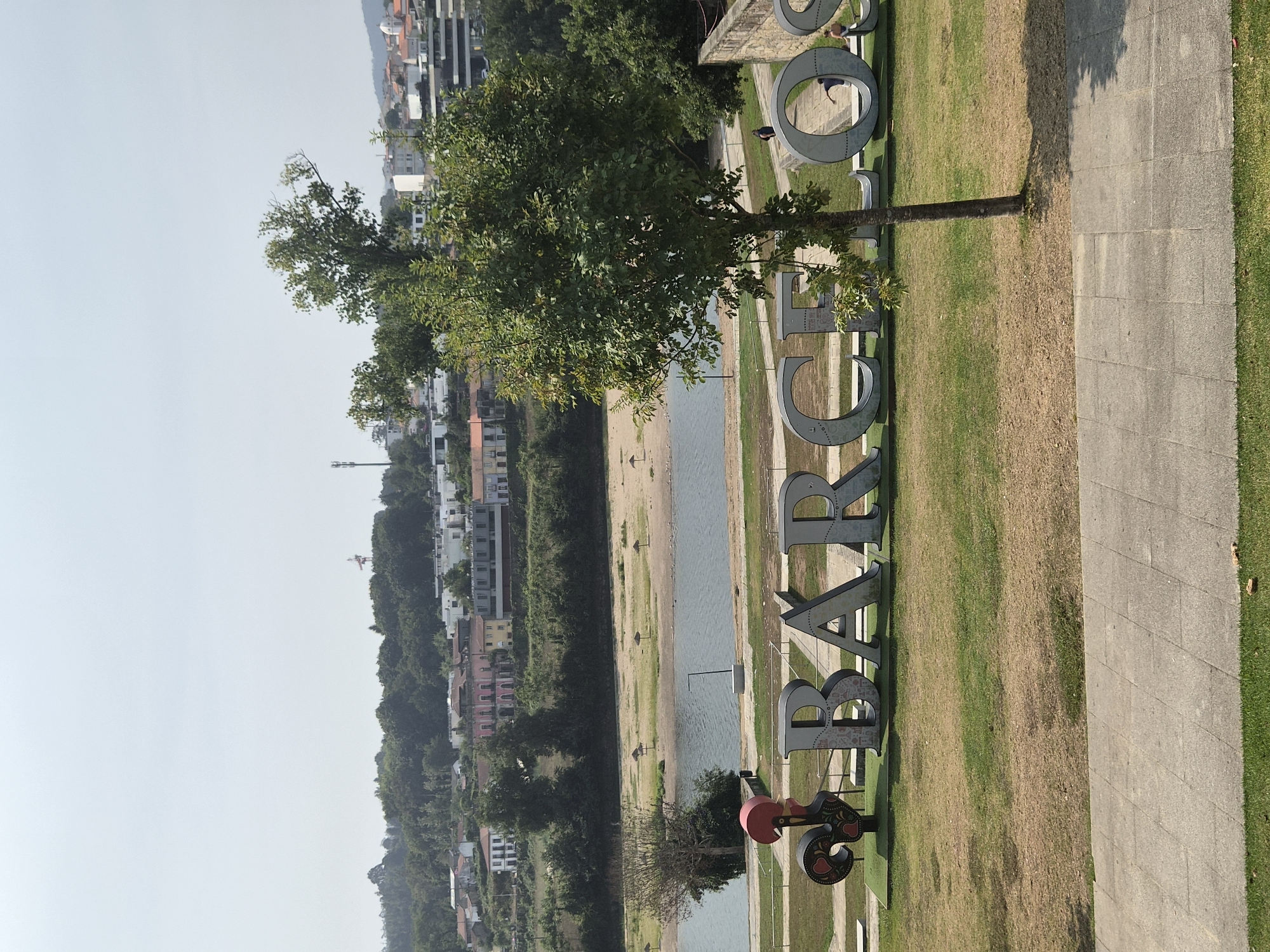 The image captures a sunny outdoor scene in Barcelos, Portugal. Dominating the foreground are large, three-dimensional metal letters spelling out "BARCELOS." To the left of the letters, a colorful rooster statue, a symbol of Barcelos, stands prominently. Behind the letters and statue, a body of water, likely a river, is visible, with a sandy beach and several small umbrellas on its bank. Further in the background, a dense green landscape of trees and vegetation extends up a gentle slope, culminating in a town with buildings of various architectural styles, including some with red-tiled roofs. The sky is a clear, pale blue, indicating fair weather. A few individuals are present in the scene, including a person walking behind the letters and two people near the riverbank. A slender tree stands in front of the letters, partially obscuring them. The foreground also features a paved walkway and a grassy area.