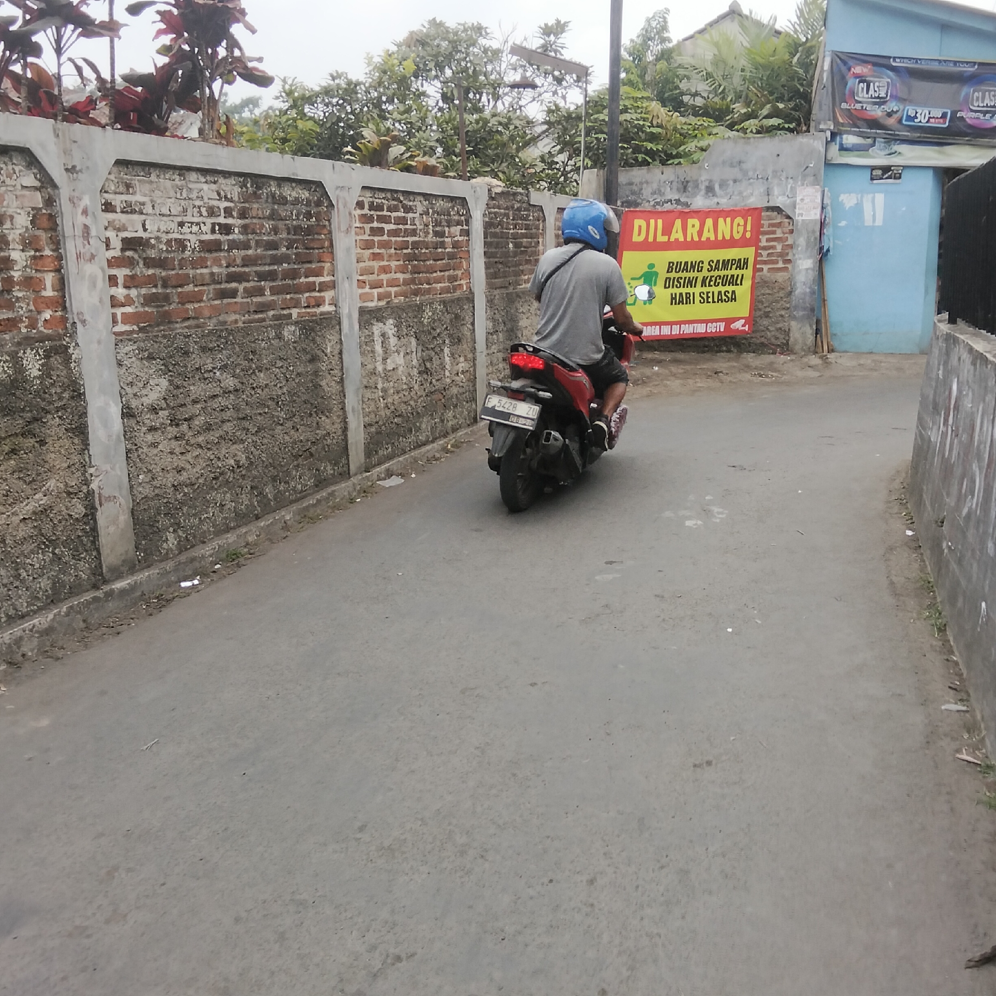 In an outdoor, urban environment, a person wearing a blue helmet and a gray shirt is riding a red motorcycle on a paved road. The motorcycle has a license plate that reads "F 5428 ZU". A red and yellow sign is attached to a brick wall behind the motorcycle, displaying text in Indonesian. The sign reads "DILARANG!" which means "PROHIBITED!" and below that, "BUANG SAMPAH DISINI KECUALI HARI SELASA" which means "DUMP TRASH HERE EXCEPT ON TUESDAY". The sign also shows an illustration of a person throwing trash into a bin, with a motorcycle beside it. Further to the right, a blue building is visible with advertisements on its side. The ground is a gray asphalt road with some debris scattered on it. A concrete wall, possibly a boundary, runs along the left side of the road. Trees and foliage are visible in the background, along with a light pole.