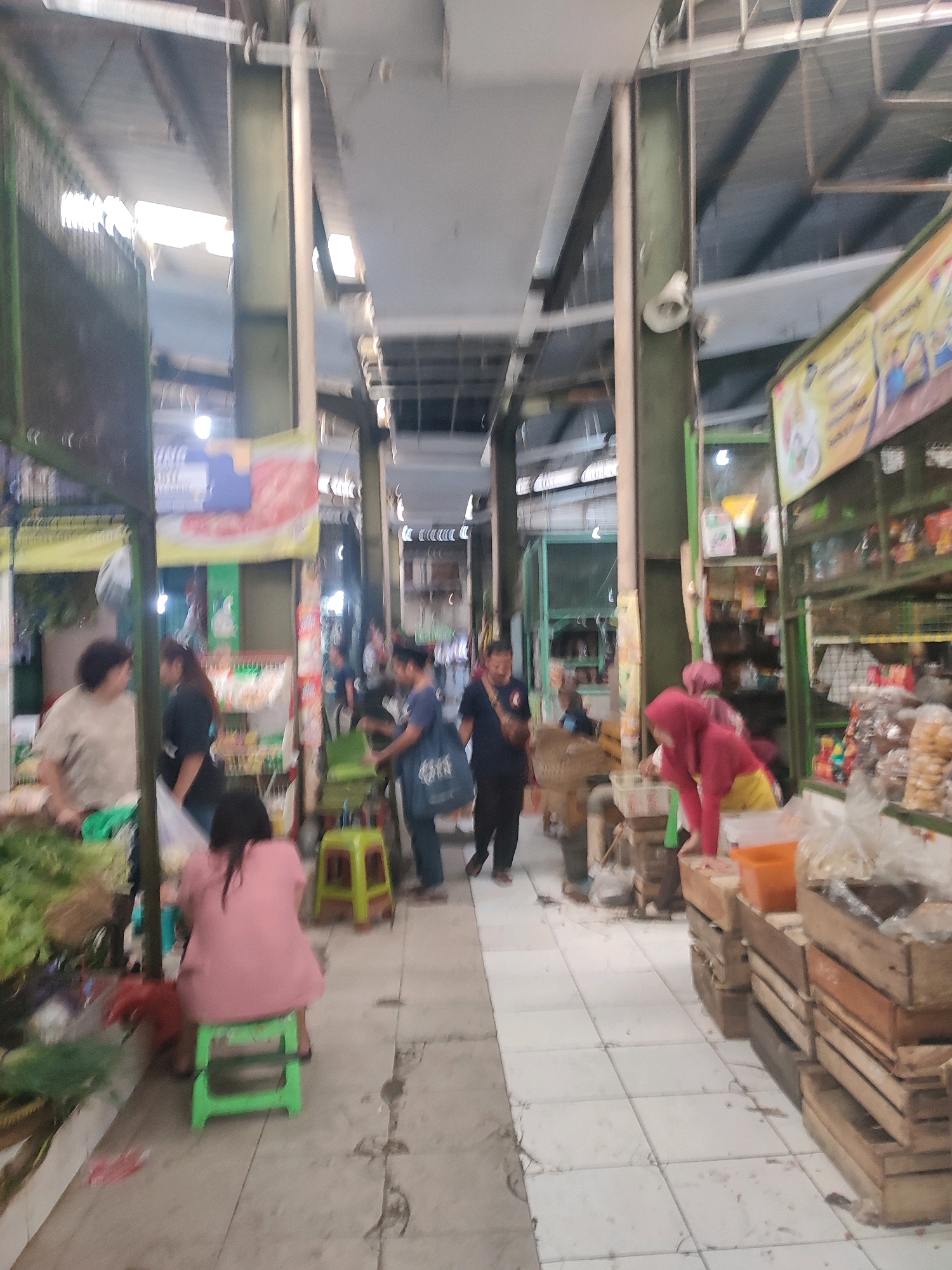 The image depicts a busy scene inside a market, likely in Yogyakarta, Indonesia. A long, covered aisle is lined with stalls on either side, showcasing various goods. People are seen browsing and interacting with vendors. The architecture features metal support beams and a corrugated metal roof, with natural light filtering in from openings.
On the left side of the aisle, a woman in a beige top is attending to a stall filled with green vegetables. Further down, a woman in a pink top is seated on a green stool, facing away from the camera. Other individuals, blurred due to motion, are also visible.
On the right side, a stall is laden with packaged goods, snacks, and other food items displayed on shelves and in crates. A person in a pink hijab and yellow pants is seen leaning over a stall, possibly arranging items or making a purchase. Another person in a blue shirt is visible in the background, near a stall with produce.
The floor is tiled, with some areas appearing worn. The overall atmosphere suggests a vibrant and active marketplace, typical of urban centers in Indonesia. The presence of various food items and the bustling activity point towards a local market where daily necessities and goods are traded.
