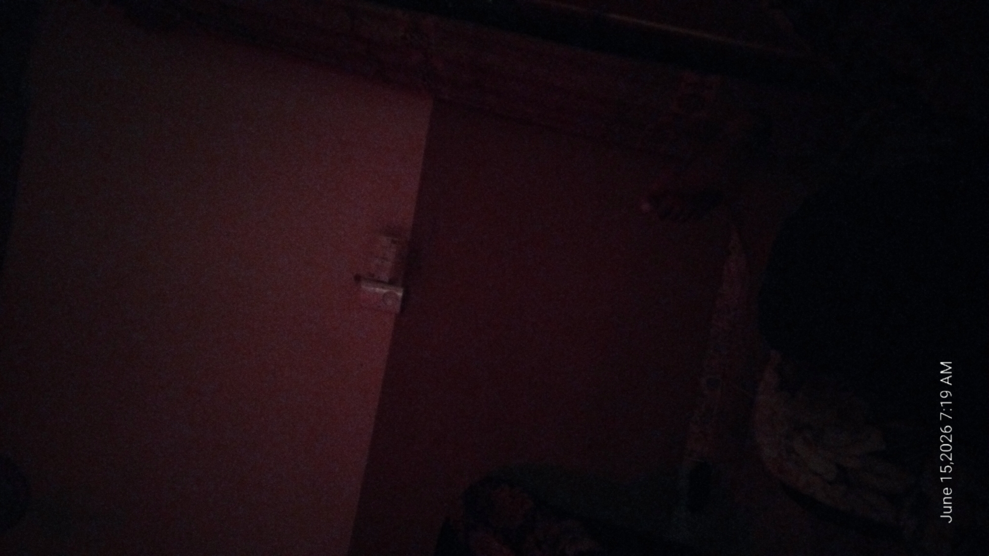The image shows a dimly lit interior scene. In the upper portion of the image, a light switch is visible on the wall. The lower portion of the image is largely dark, but a person's body, possibly lying down, is partially visible, covered by a patterned textile. A date and time stamp in the bottom left corner reads "June 15, 2026 7:19 AM". The location is stated to be in the city of Howayi, Nigeria.