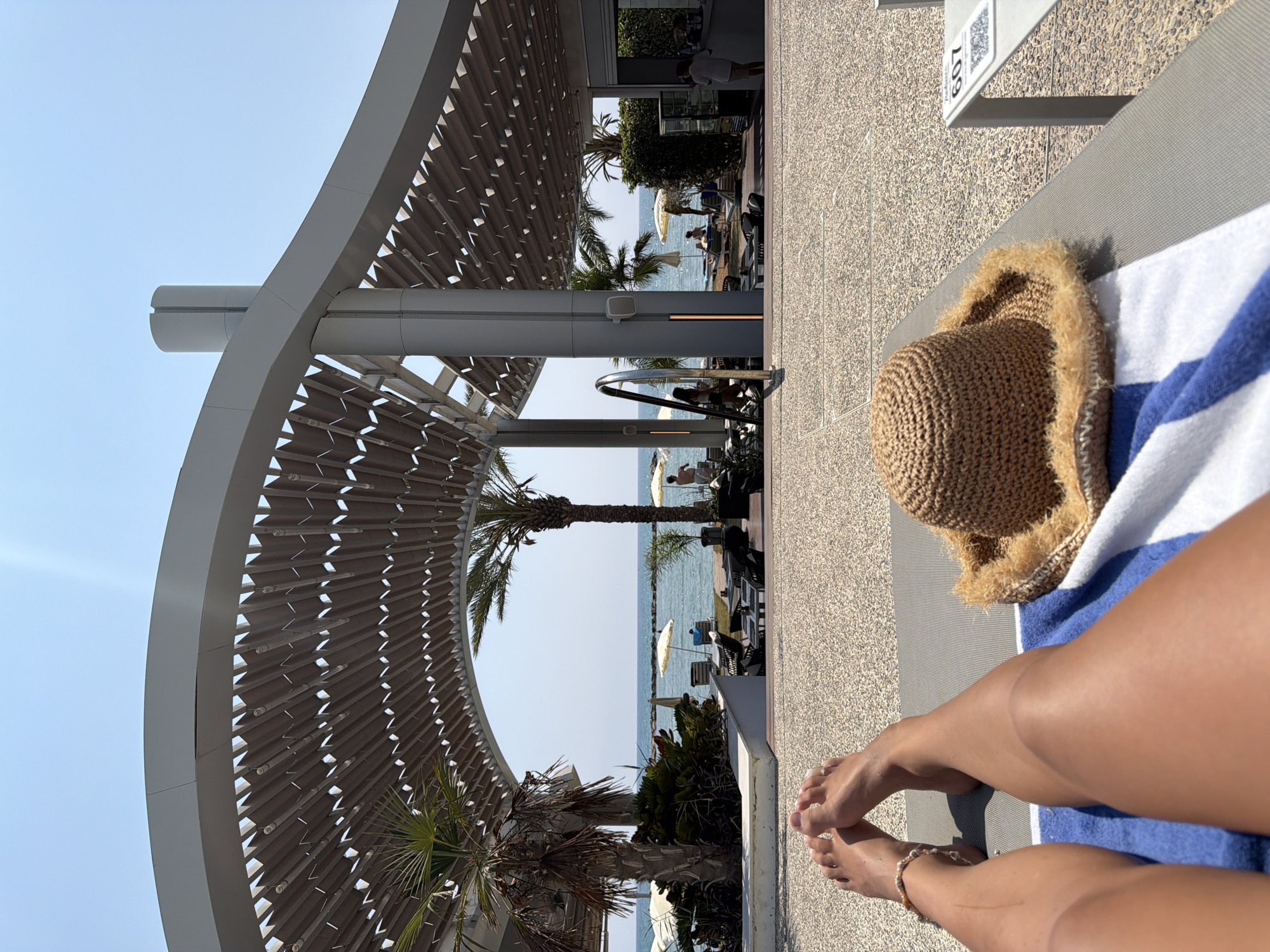 The image depicts a sun-drenched scene at a beachfront establishment in Oroklini, Cyprus. In the foreground, a person's legs and feet are visible, resting on a lounge chair covered with a blue and white striped towel. A straw hat with a frayed brim sits on the towel next to the person's legs. The lounge chair is situated on a textured, pebble-like ground.
Beyond the lounge chair, the scene opens up to reveal a view of the sea. A modern, open-air structure with a curved, slatted roof provides shade. Palm trees are present in the middle ground, one on the left and another to the right, framing the view of the beach and ocean. Several beach umbrellas and lounge chairs are scattered on the sand. People are visible in the distance, some walking on the beach and others sitting at tables outside a building that appears to be a bar or restaurant. The sky is clear and blue, indicating a sunny day. A QR code with the number 607 is visible on a table to the right of the frame.