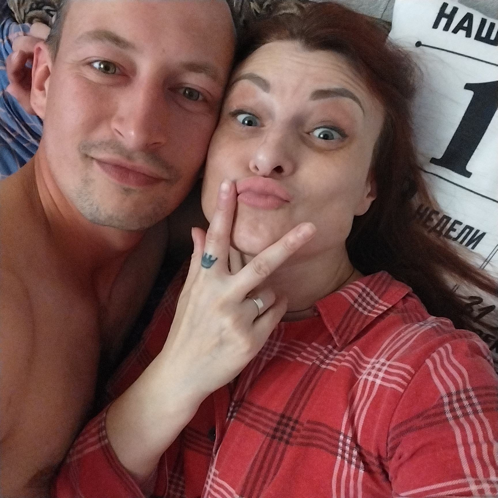 A close-up, overhead selfie shows a man and a woman lying down and posing for the camera. The man's head is on the left, and he is looking slightly away from the camera with a faint smile. His skin is tanned, and he has light stubble on his chin and upper lip. The woman is on the right, with her eyes wide open and her lips pursed as if blowing a kiss. Her hair is dark brown and falls around her face. She is wearing a red plaid shirt. Her left hand is raised to her face, with her index finger touching her lips. She has a small tattoo of a crown on her left middle finger and a silver ring on her ring finger. Behind the couple, a pillow with text is visible. The lighting appears to be natural, possibly from a window. The background is indistinct, suggesting an indoor setting.