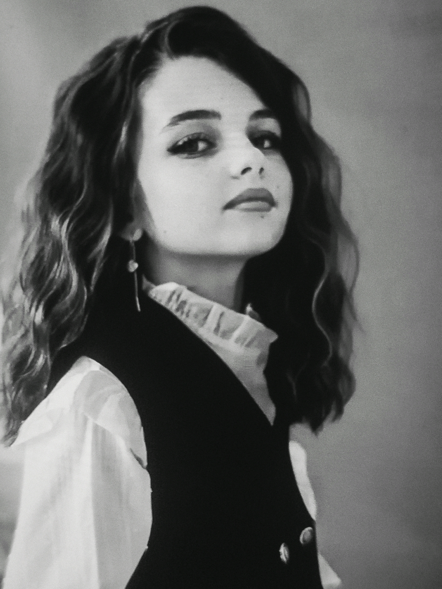 A black and white medium shot captures a young woman with dark, wavy hair styled in soft curls. She is positioned in a three-quarter profile, looking towards the viewer with a slight tilt of her head. Her makeup is noticeable, with dark eyeliner emphasizing her eyes and lipstick coloring her lips. She wears a white collared shirt with a ruffled neckline peeking out from under a dark vest. A long, dangling earring with a circular stud is visible on her left ear. The background is a plain, muted gray, offering no discernible details about the environment or location.