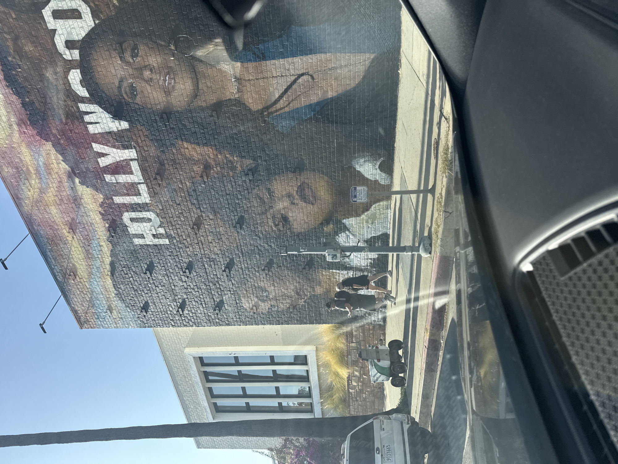 The image captures a view from inside a vehicle, focusing on a large mural on a brick wall. The mural features two prominent black figures and the word "HOLLYWOOD." The location is likely within the city of Los Angeles, United States of America, given the mural's subject and the presence of the "HOLLYWOOD" inscription. The mural's imagery includes what appears to be stylized depictions of figures against a background suggesting an urban or sky scene. Several birds are depicted perched on the brick wall.
On the sidewalk beside the mural, two people are seen jogging. A small delivery robot is near the curb. A white van is parked further down the street. Streetlights and a charging station sign are positioned along the sidewalk. The overall scene presents a blend of art, urban life, and street activity.