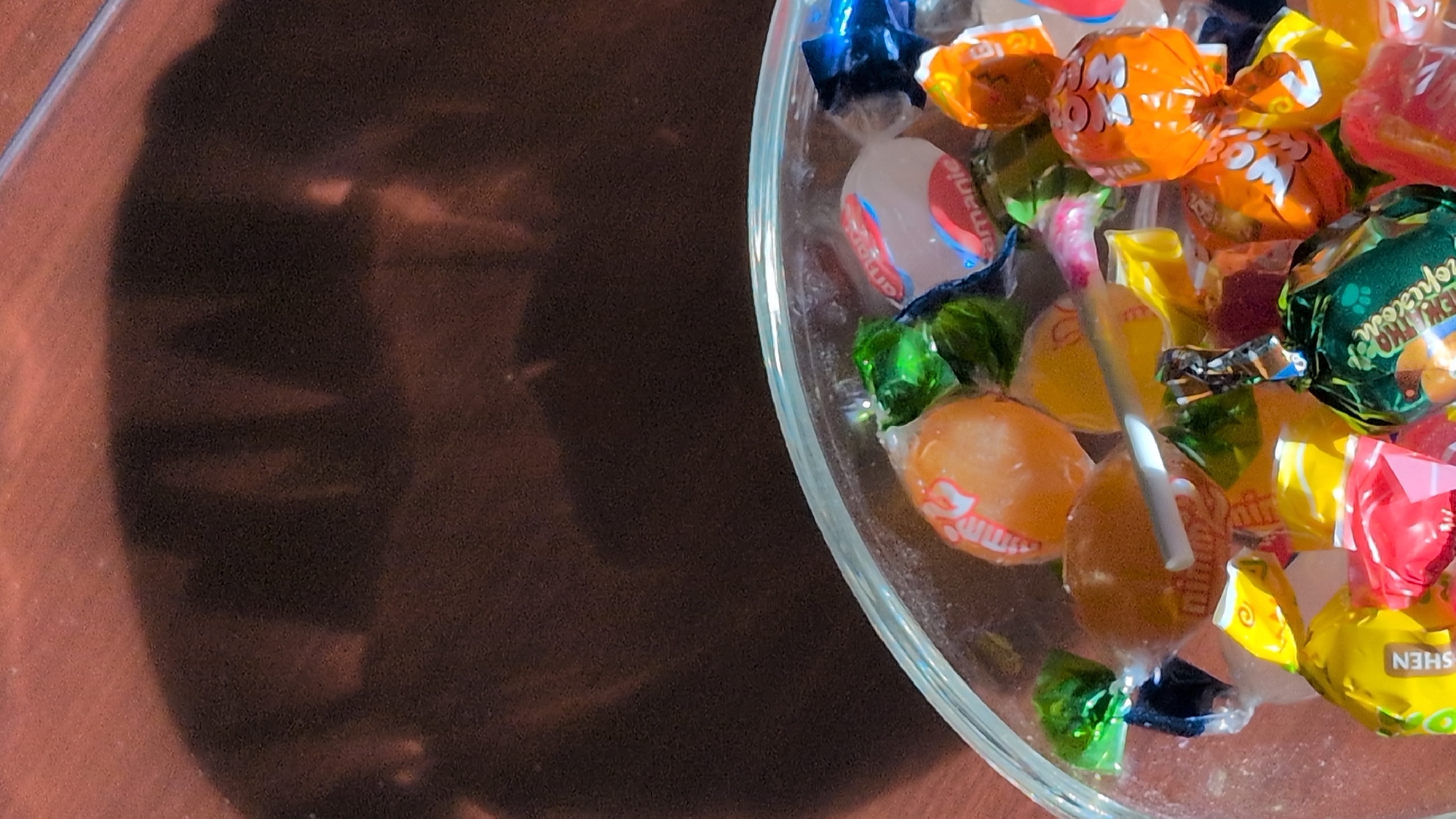 The image displays a close-up view of a clear glass bowl filled with an assortment of individually wrapped candies. The candies are of various colors, including orange, yellow, green, blue, and red, and feature different wrapper designs and text. Some wrappers show visible text such as "Mini", "kamani", and "Shen". A thin, silver stick-like object is also visible among the candies. The bowl is placed on a brown surface, likely a wooden table, which is illuminated by a light source casting a large, dark shadow on the upper portion of the image. The shadow appears to be cast by an object or possibly a person, with indistinct shapes and contours. The scene is set indoors, with the wooden surface and lighting suggesting a domestic or indoor setting. There is no discernible indication of the city of Rastenberg, Germany, in the image itself, and therefore no context for that location can be provided based on the visual information.
