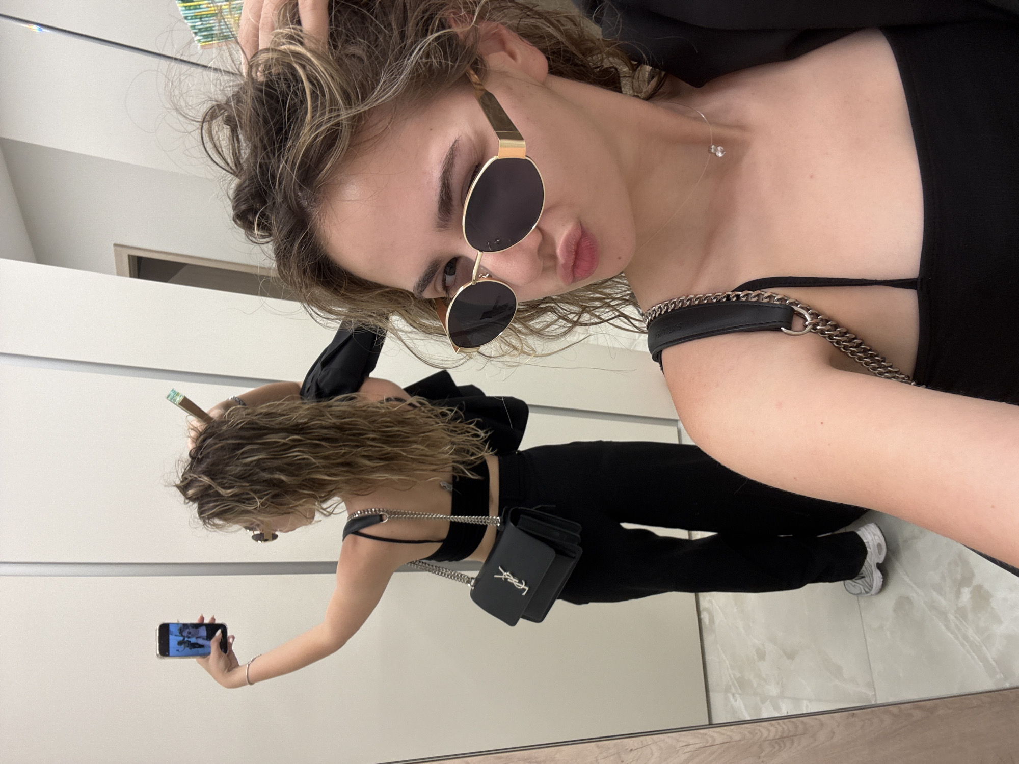 A young adult female is depicted taking a mirror selfie in a well-lit indoor environment. The setting features large, seamless white wall panels or mirrors, and a light-colored, possibly marble or polished tile floor with subtle grey veining. The specific location within Sofia, Bulgaria, cannot be definitively identified from the visual information presented; however, the setting appears to be a modern indoor space, likely a retail changing room or a well-appointed hallway.
The woman, with medium-length wavy brown hair highlighted with blonde streaks, is positioned close to the camera, capturing a direct shot of her upper body and a full reflection of her from the back. She wears oval, gold-framed sunglasses with dark lenses and puckers her lips in a 'kiss' pose, looking directly at the camera. Her left hand is touching her hair near the scalp. She is wearing a black strappy crop top and a delicate transparent necklace with a single clear gem pendant. A black blazer or jacket is draped over her left shoulder.
In the mirror reflection, her full figure is visible from the back. She is holding a smartphone (displaying her own image) in her raised right hand, adorned with a silver-toned bracelet. A black Yves Saint Laurent (YSL) Sunset bag with a silver chain strap is slung over her left shoulder. She is also wearing black high-waisted, wide-leg pants and grey and white athletic sneakers. A slender object with a greenish tip, possibly a vape pen, is visible near her reflected left hand/hair. The reflected background includes a light brown wooden doorframe with a white door, and above her head, a section of a multi-colored (green, yellow, blue) pattern, possibly shelving or merchandise.