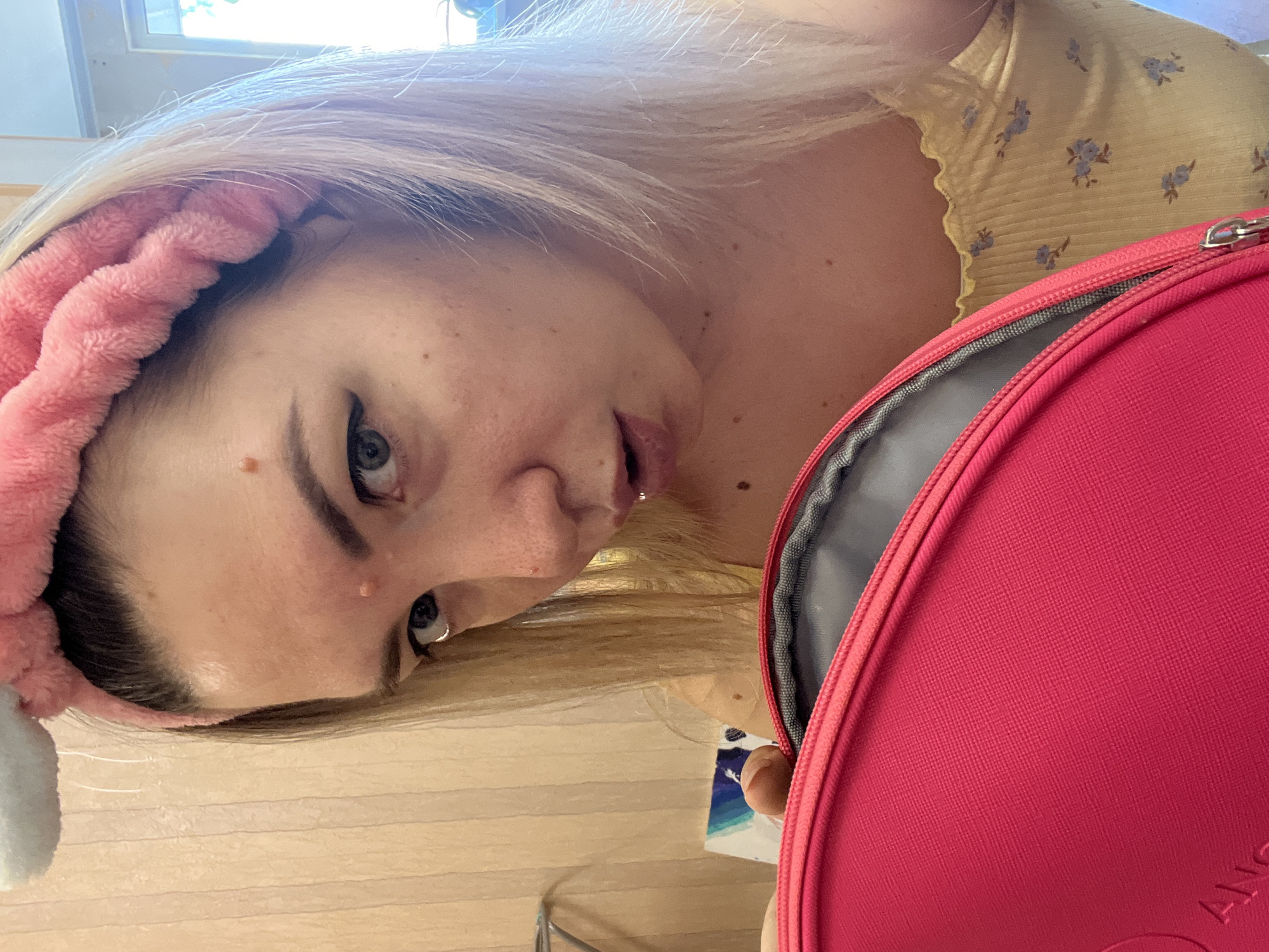 The media file is a close-up, low-angle selfie of a young woman with blonde hair. She is wearing a pink fluffy headband. Her head is tilted, and she is looking towards the camera with one eye visible. She appears to be wearing makeup, with noticeable eyeliner. Her skin has freckles and a few small blemishes. She is wearing a yellow top with a small floral pattern. To the right of the frame, a bright pink object, likely a bag or case, is visible, partially open and revealing a gray interior. The background consists of what appears to be a wooden surface, possibly a table or floor, and a window with light streaming through. The overall mood of the image is casual and personal. There is no visible text. The location context of Sochi, Russia does not provide any specific cues within the image itself.