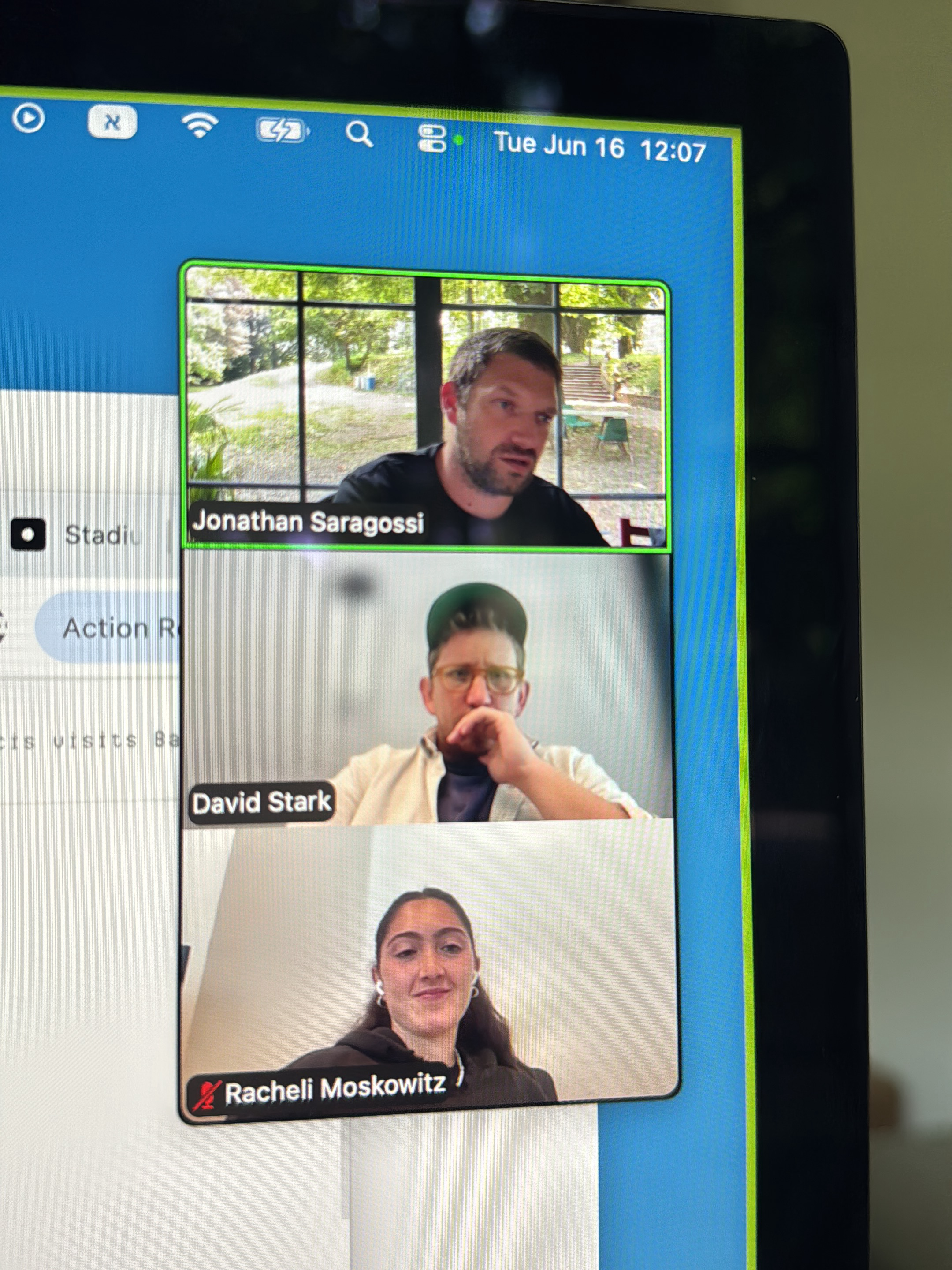 The image shows a computer screen displaying a video conference call. Three individuals are visible in separate video windows. The top window shows Jonathan Saragossi, a man with short brown hair and a beard, wearing a black t-shirt. He is speaking and looking to his right. Behind him, a window reveals an outdoor scene with trees and stairs. The middle window shows David Stark, a man wearing glasses and a green baseball cap. He has his hand on his chin and appears to be listening. The bottom window features Racheli Moskowitz, a woman with dark hair pulled back, wearing a black hoodie and white earbuds. She is smiling. Below her name, a red icon indicates her microphone is muted.
The top portion of the computer screen shows the operating system's status bar with icons for Wi-Fi, battery, and a search function. The date and time "Tue Jun 16 12:07" are also visible. To the left of the video windows, a sidebar with text such as "Stadiu" and "Action R" suggests a tabbed interface or application menu. The overall setting appears to be indoors, with the video conference being the primary activity. The lighting suggests it is daytime.