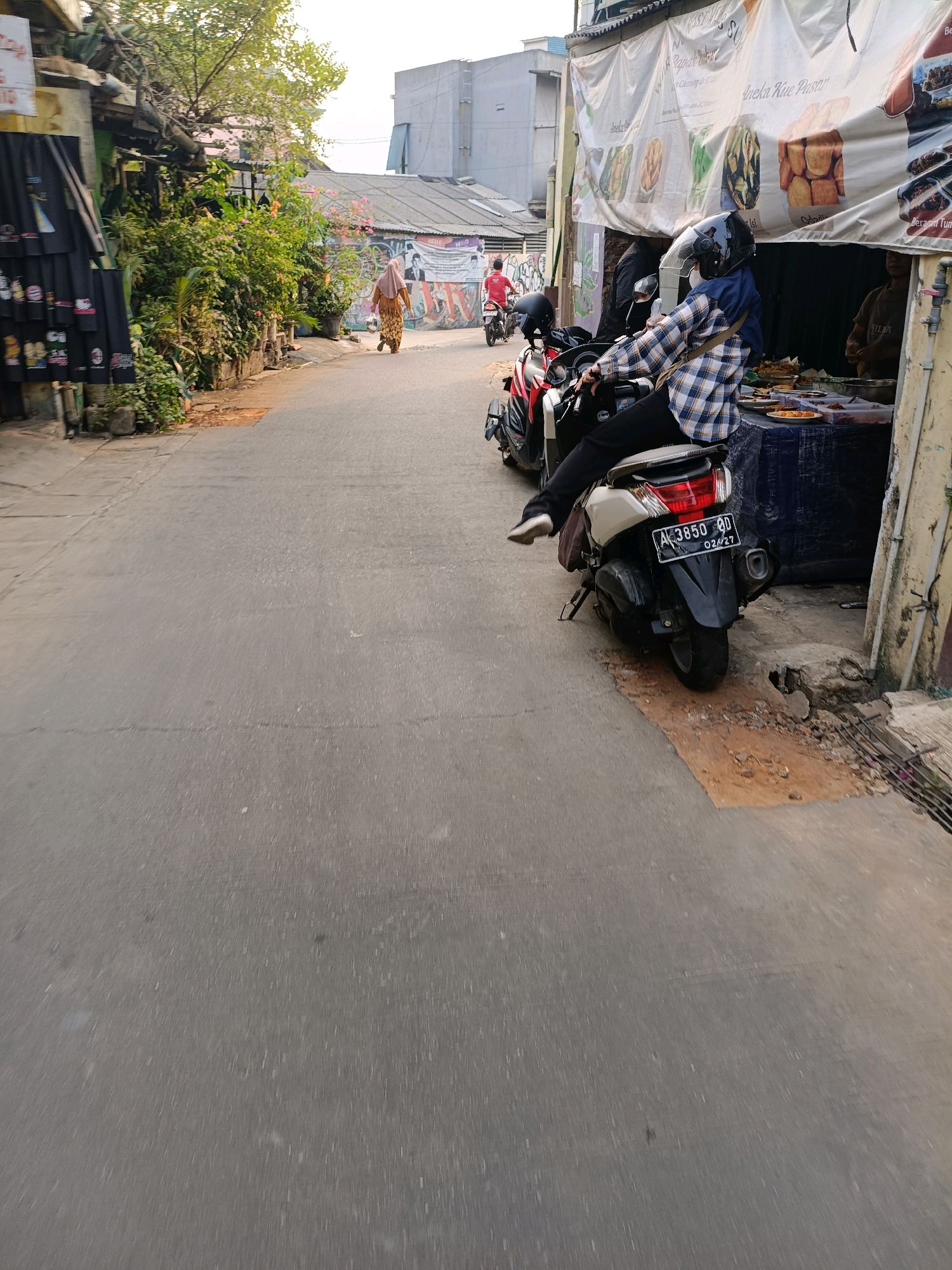 In Tangerang, Indonesia, a street scene unfolds with a focus on a food stall and passing pedestrians and vehicles. The stall, draped with a large banner displaying images of food items and text in Indonesian, is situated on the right side of the frame. A woman wearing a blue hijab and a checkered shirt is seated on a white scooter parked in front of the stall. She has her right leg extended, and her left leg is bent. Another scooter is parked behind hers. To the left of the food stall, a person in a brown shirt is visible. In the background, a woman in a long brown garment walks away from the camera, and a person on a red scooter is seen further down the street. The street is paved with asphalt, and the surrounding environment features buildings with various roofs, lush greenery, and some signage. The lighting suggests it is daytime.