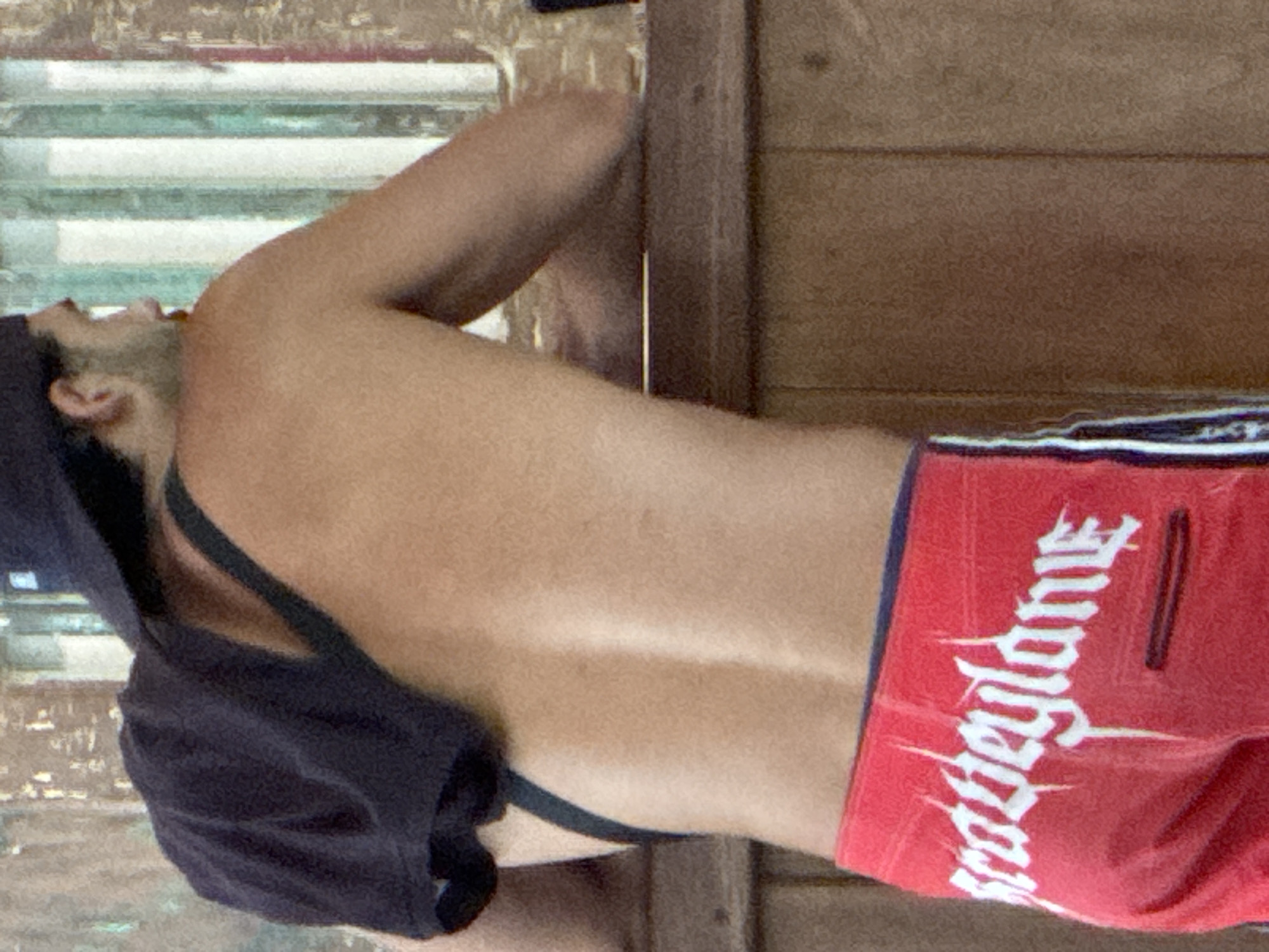 The image shows a male individual, presumably in Kuta Selatan, Indonesia, captured from a back-side perspective. The subject is resting or lying down during the daytime, with his head tilted back and looking upwards. He has short dark hair and some facial hair. His upper body is bare, revealing his back and shoulder, suggesting he is shirtless or wearing minimal upper body clothing. A dark strap, possibly from a tank top, swimsuit, or backpack, is visible over his shoulder.
He is wearing red shorts that feature prominent white, stylized text on the left side, which appears to read "Wadeyane" in a gothic-style font. The background directly behind him consists of light-colored horizontal slats or blinds, while to his right, there is a darker wooden panel or planked surface. The lighting indicates it is daytime, casting a soft illumination on the scene. No specific activities beyond resting are discernible.