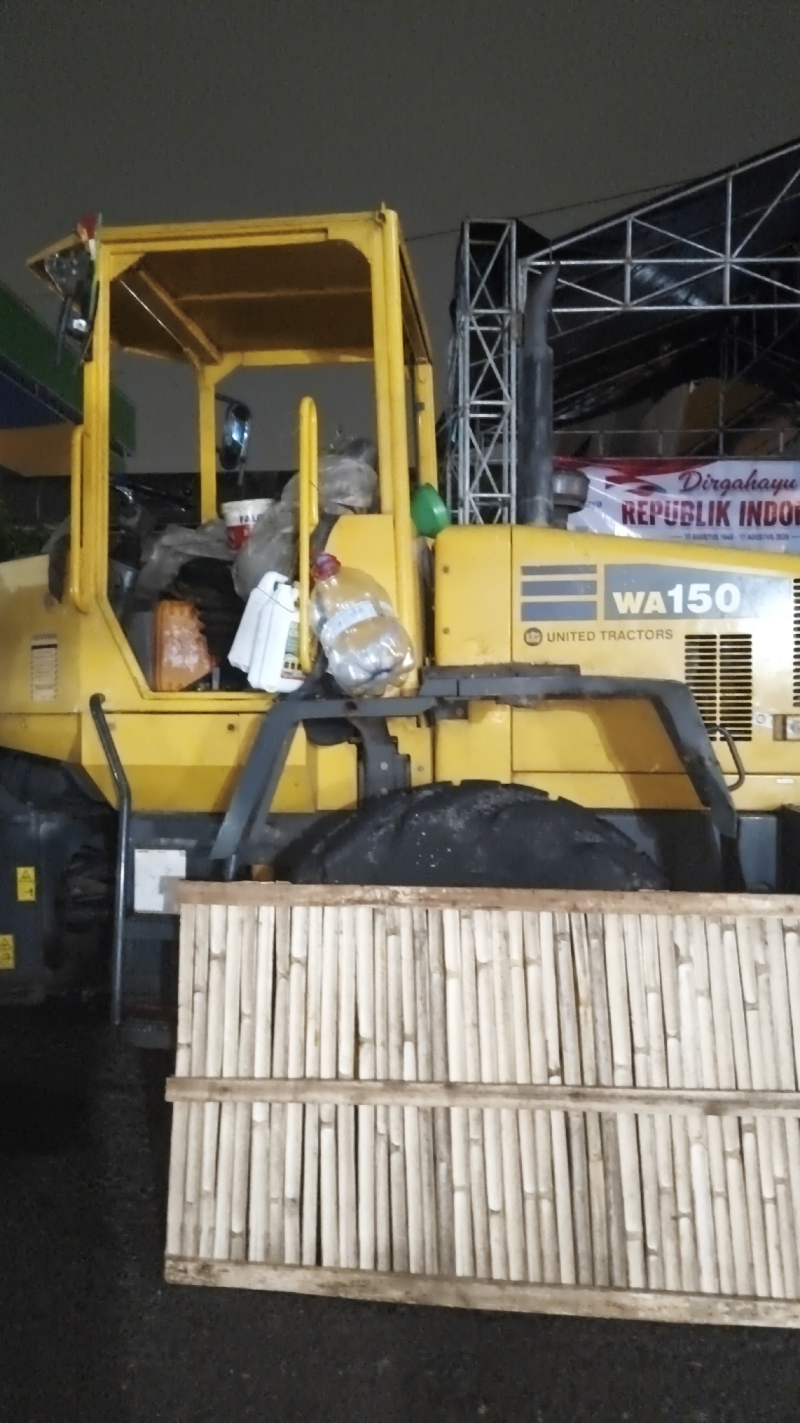 A yellow Komatsu WA150 wheel loader is parked on an asphalt surface at night. The wheel loader has a visible cab with an interior that appears to contain plastic sheeting and several containers. A large, clear plastic bottle with a red cap is perched on top of a white container within the cab. To the right of the wheel loader, a banner displaying "Dirgahayu REPUBLIK INDONESIA" is partially visible, suggesting the location is in Indonesia. In front of the wheel loader is a barrier constructed from bamboo poles bound together with horizontal wooden planks. The background features a dark, possibly cloudy sky and metal scaffolding, indicating an outdoor event or construction site. The scene is lit by artificial light, creating shadows and highlighting the yellow machinery. There are no people visible in the image. The scene appears to be located in South Jakarta, Indonesia, as indicated by the banner.