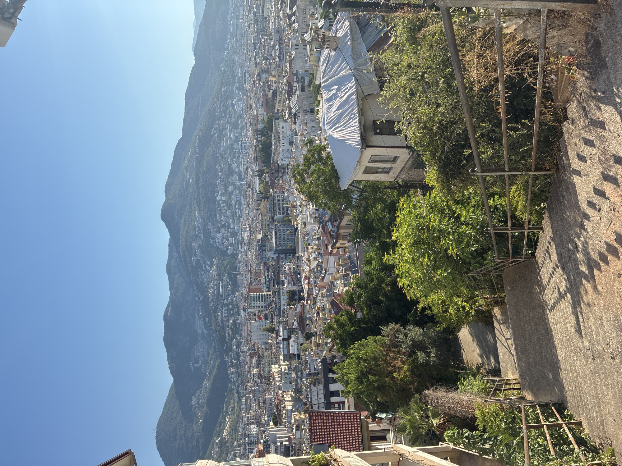 The image shows a downward view of a concrete staircase with metal railings on either side, leading into a dense green foliage area. Beyond the stairs and foliage, a sprawling city with numerous buildings and a distant mountainous backdrop can be seen under a clear blue sky. The city appears to be Alanya, Turkey, as indicated by a sign on the mountainside in the upper center portion of the image. The architecture of the buildings in the city suggests a mix of residential and commercial structures, many with flat roofs topped with solar water heaters. A distinct building with a white tarp covering its roof is situated on the right side, amidst the greenery. The overall scene captures a sunny day in a coastal city, with a significant contrast between the natural environment of the foreground and the urban landscape in the background.
