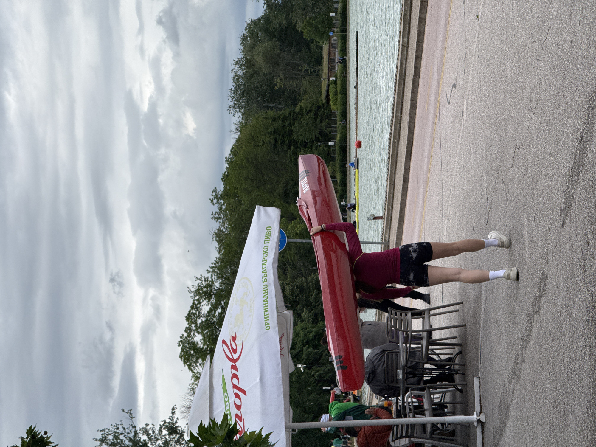 A person is carrying a red kayak overhead, near a body of water, and tables and chairs are visible in the foreground. A large white umbrella with green writing on it, which reads "Каменица Оригинално Българско Пиво" and an image of a wheat stalk, partially obscures the view on the left side of the frame. The writing indicates it is advertising Kamenitza, a Bulgarian beer. The body of water in the background looks like a rowing channel; the scene is likely in Plovdiv, Bulgaria, where the Plovdiv Rowing Canal is located. There are trees and vegetation in the background along the edge of the body of water. The sky is overcast with grey clouds.