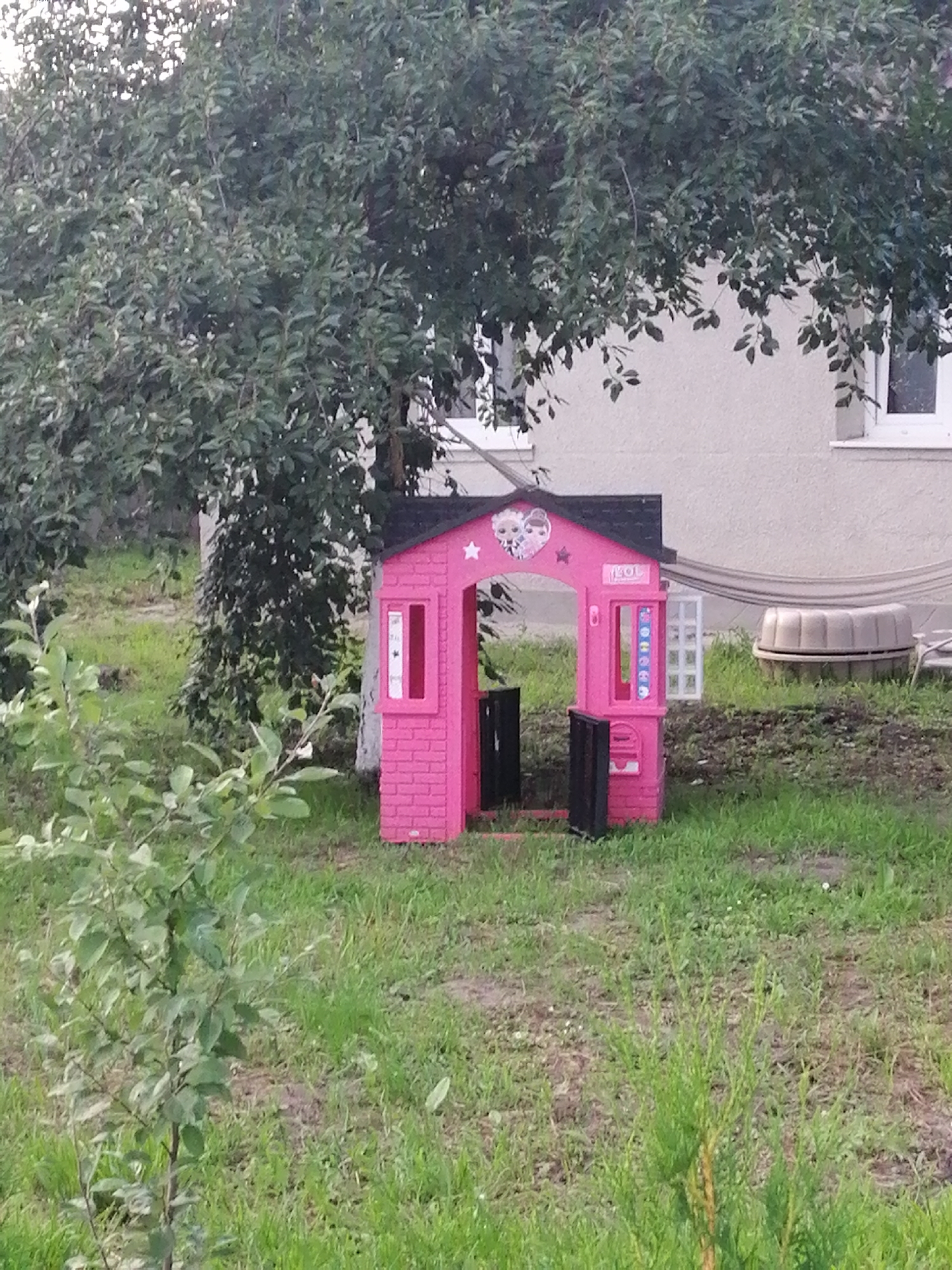 In Lanivtsi, Ukraine, a pink plastic playhouse with a black roof stands on a grassy lawn in front of a light-colored building. The playhouse has two windows on the front, one with a white frame and vertical bars, and an open doorway with black doors. A decal with cartoon characters and stars is visible on the front of the playhouse. To the right of the playhouse, there is a white window on the building and a beige sandpit with a ribbed pattern. A hammock is draped over the sandpit. A large tree with green leaves shades the playhouse and the building. A small, leafy sapling is in the foreground to the left, and a small evergreen tree is in the foreground to the right. The scene appears to be a backyard or garden area.