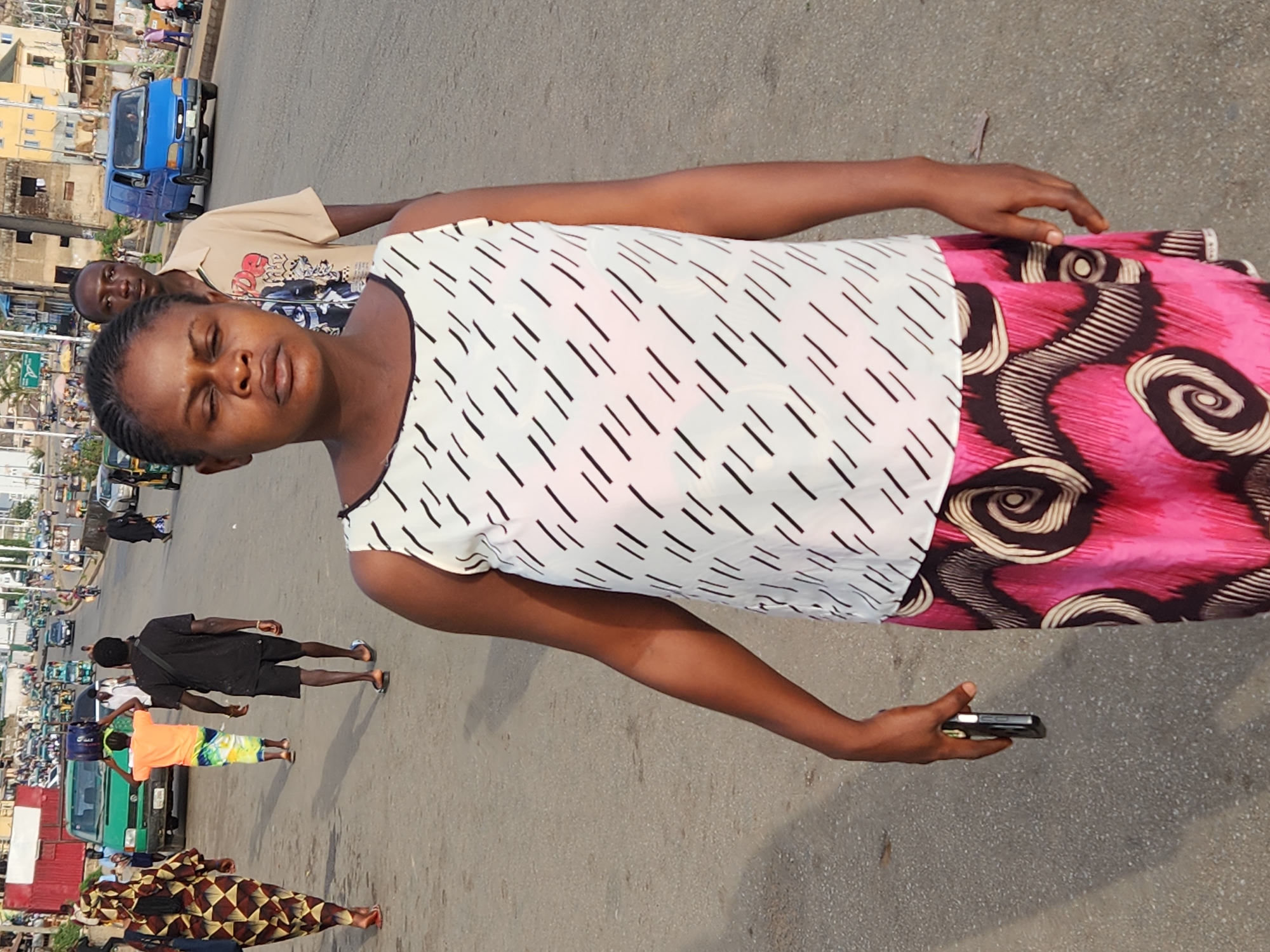 A street scene in Jos, Nigeria, features a woman as the primary subject. She has dark skin, braided hair, and is wearing a white top with black stripes and a pink patterned skirt. She holds a phone in her right hand. A man stands behind her, wearing a light-colored shirt. Pedestrians walk on the street, and vehicles are visible, including a blue van, a green vehicle, and what appears to be motorcycles or tricycles further down the road. Buildings line the street in the background. The surface beneath the woman is paved, likely asphalt or concrete.