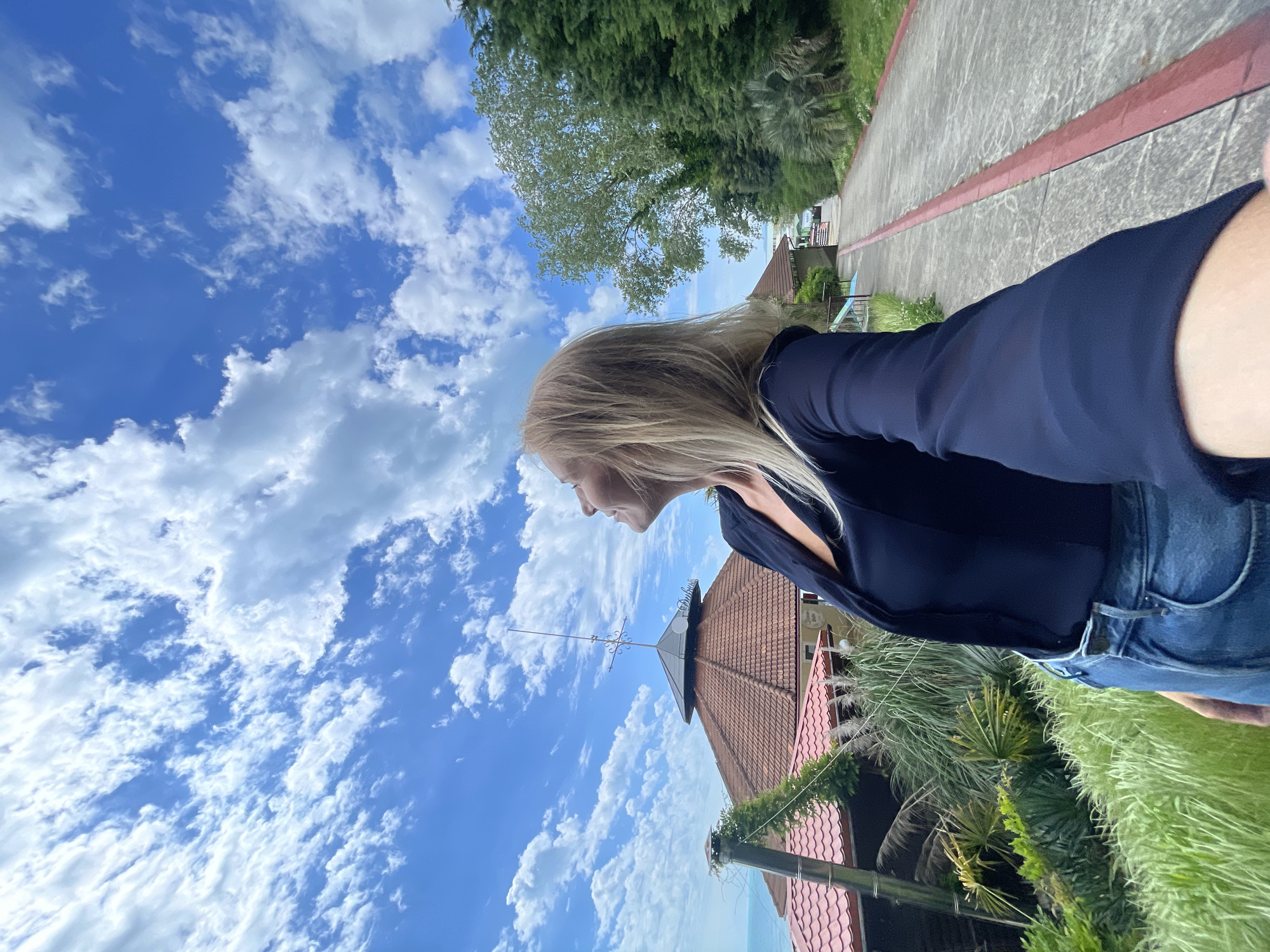 A woman with blonde hair, wearing a dark blue blouse and jeans, is seen in an outdoor setting. The background includes a bright blue sky with white clouds, a paved walkway lined with trees, buildings with red-tiled roofs, and lush greenery. The buildings and foliage suggest a location possibly in the city of Sochi, Russia.