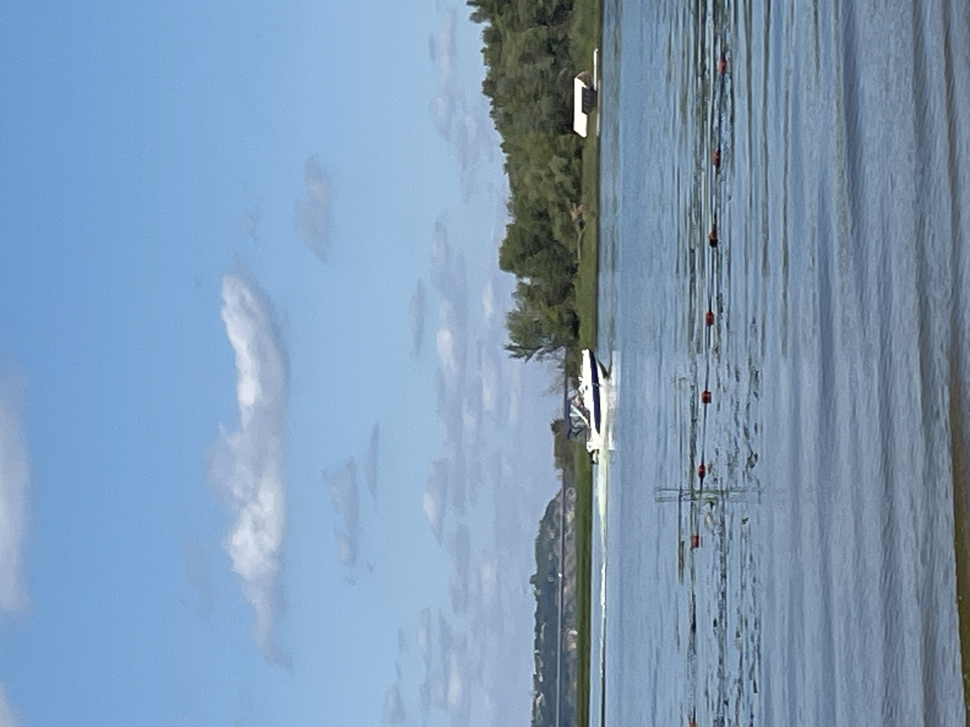 A motorboat, white with blue trim, is navigating a body of water. The boat is creating a wake. In the background, there is a tree-lined shore with a small white building on a dock. A bridge is visible in the far distance. The sky is blue with scattered white clouds. The water in the foreground is calm and shows ripples. Red buoys are visible in the water. The scene appears to be set in Shumeika, Russia.