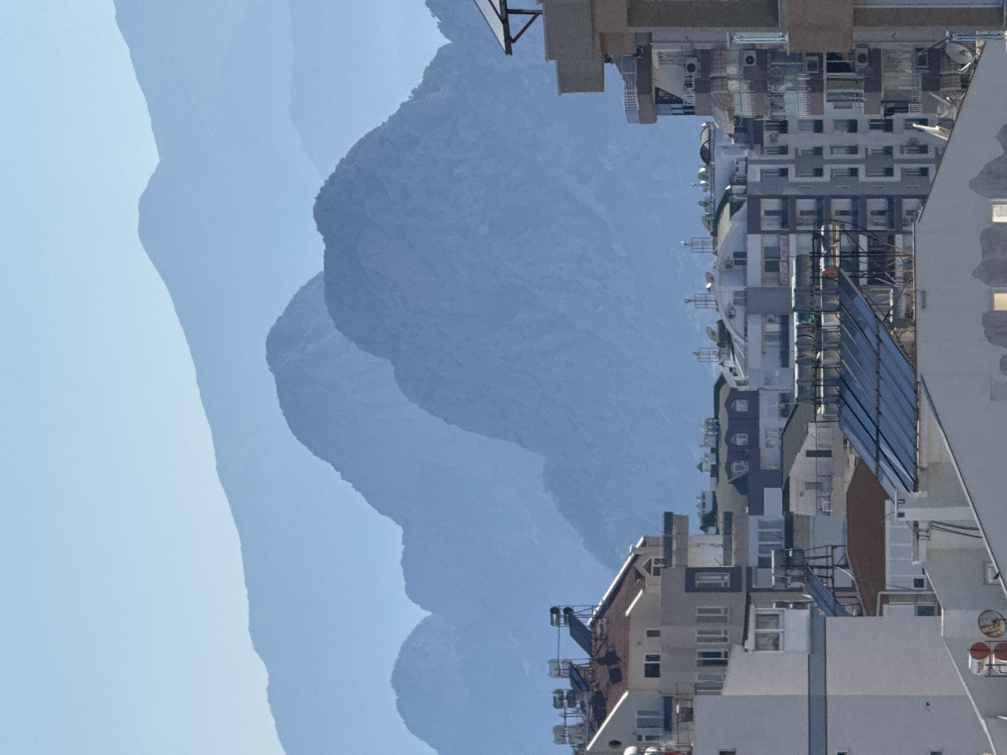 This is a vertically oriented, low-angle shot of a city skyline with mountains in the background. The city appears to be built on a hillside, with numerous multi-story buildings packed closely together. The architecture is modern, featuring a mix of white, grey, and brown facades, with many windows visible. Some buildings have balconies or external structures, and there are antennas and solar panels on some rooftops. The mountains in the background are a muted blue, suggesting they are a significant distance away and partially obscured by haze or the sky. The sky itself is a pale blue, indicating it is daytime, likely late morning or afternoon, with clear weather. There is no visible human activity or any text in the image. The scene conveys a sense of urban density against a natural backdrop, characteristic of a coastal city.