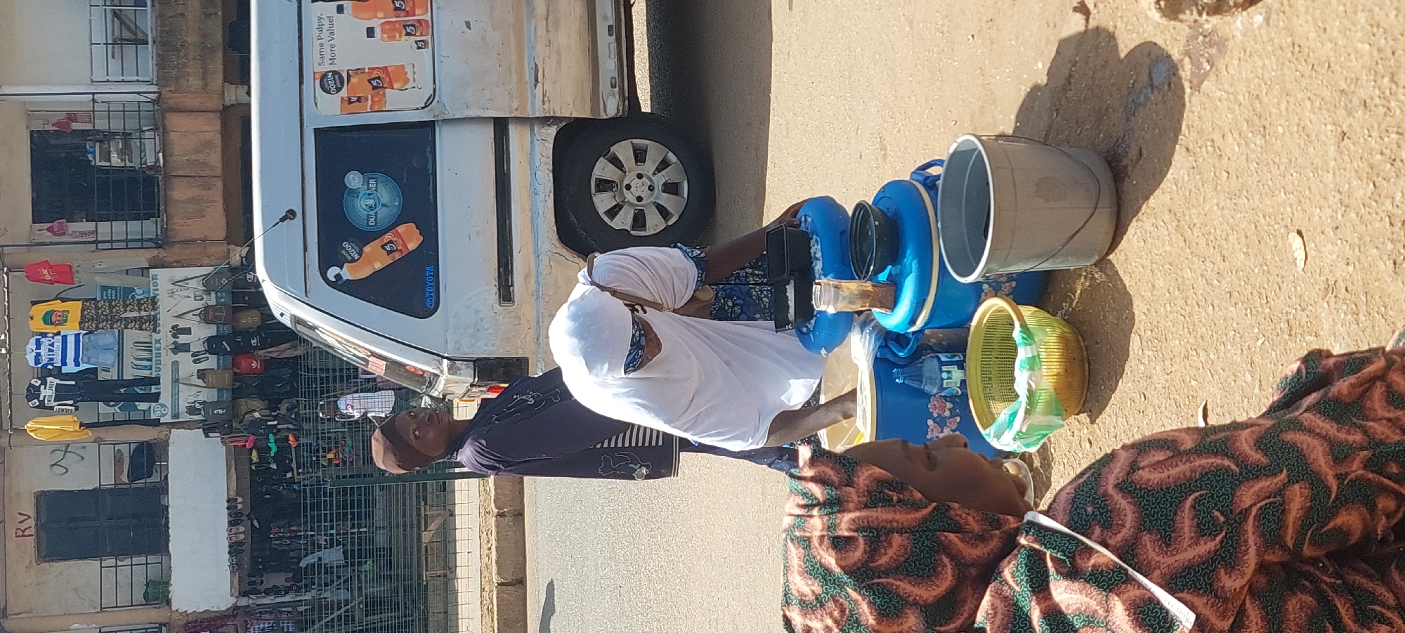 In a sunlit outdoor environment, likely a street market, two women are engaged in an activity near a white van. The woman in the foreground, on the left, wears a headscarf and a patterned fabric clothing. Her gaze is directed downwards. Behind her, another woman, wearing a white hijab and blue and white patterned clothing, is bent over a collection of blue containers. She is in the process of handling something within these containers, possibly preparing food or drinks. There are several containers, including two prominent blue ones, a yellow basket, and a grey bucket. The van, parked on the right side of the image, features advertisements and the "TOYOTA" logo. In the background, a storefront displays clothing and accessories. The ground is paved, and the overall scene suggests a commercial, busy street.
