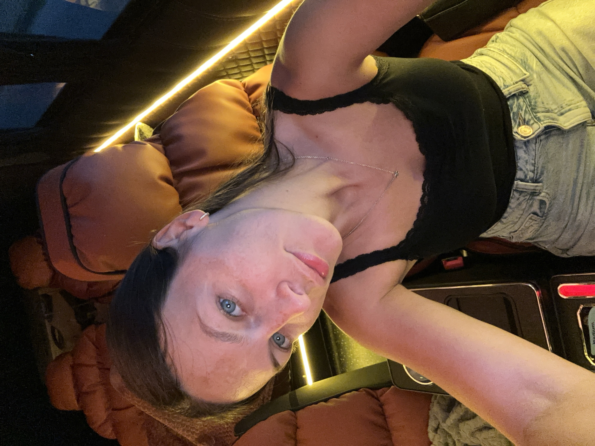 A woman is lying down in what appears to be the interior of a vehicle, possibly a car or van, indicated by the seating upholstery and the presence of a panel with electronic controls. The vehicle's interior features ambient lighting, as evidenced by the glowing linear lights. The woman is wearing a black top with lace trim and light-colored denim shorts or jeans. The image does not provide enough information to conclusively determine the location as Harsefeld, Germany. There are no visible objects or actions in the frame to determine any interactions or the environment.