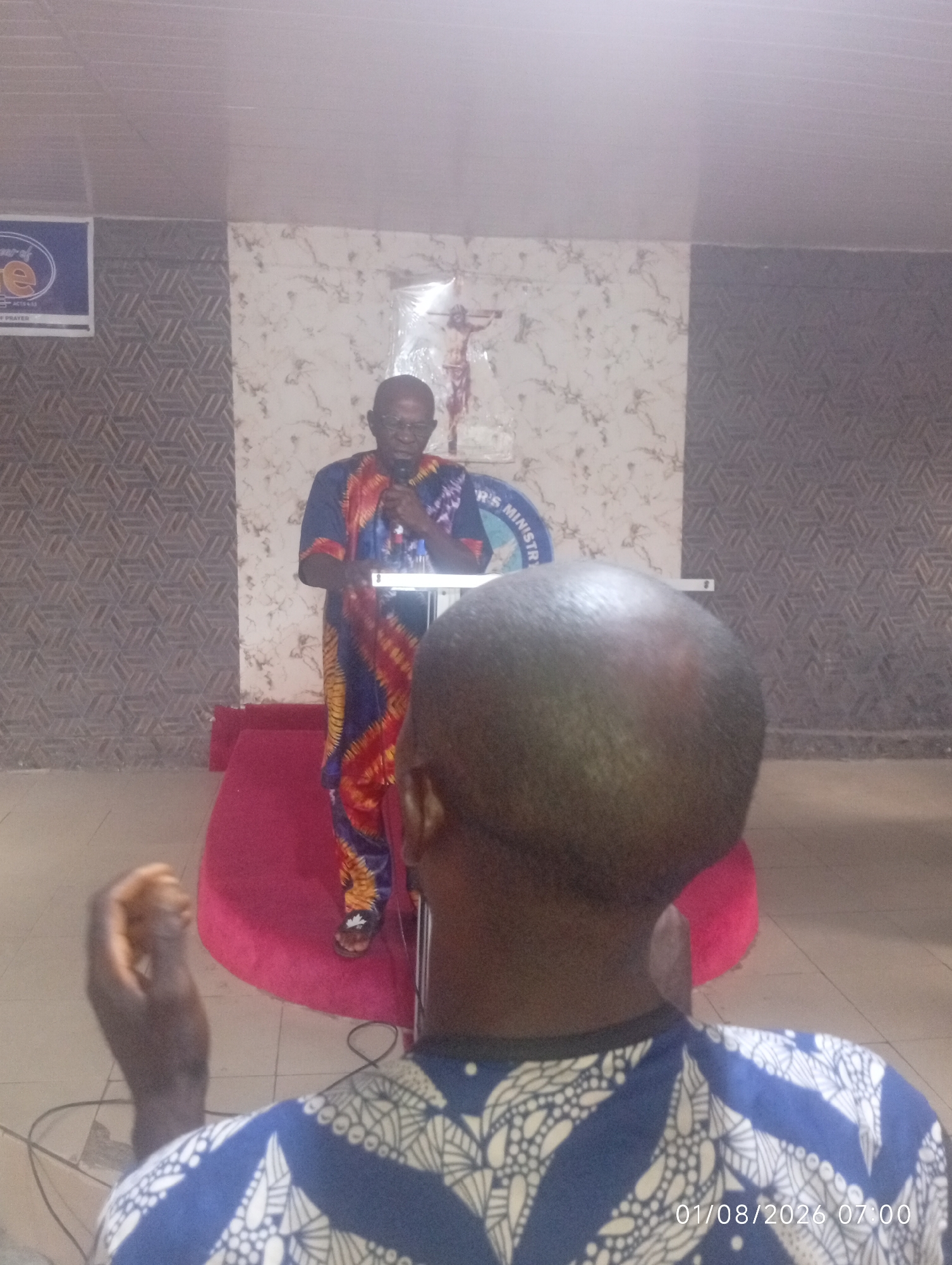 An over-the-shoulder shot from behind shows a man in a patterned blue and white shirt facing away from the camera, towards a man standing at a podium. The man at the podium is wearing a vibrant blue, yellow, and red patterned traditional outfit and glasses. He is speaking into a microphone and is positioned on a red carpeted platform. Behind him, a cross is visible. The environment appears to be an indoor space with patterned walls and a light-colored ceiling. A sign with text, including "R'S MINISTRY" and "PRAYER," is partially visible on the left. The timestamp "01/08/2026 07:00" is overlaid on the bottom right of the image. The scene is likely a church or a similar religious gathering in Lokoja, Nigeria.