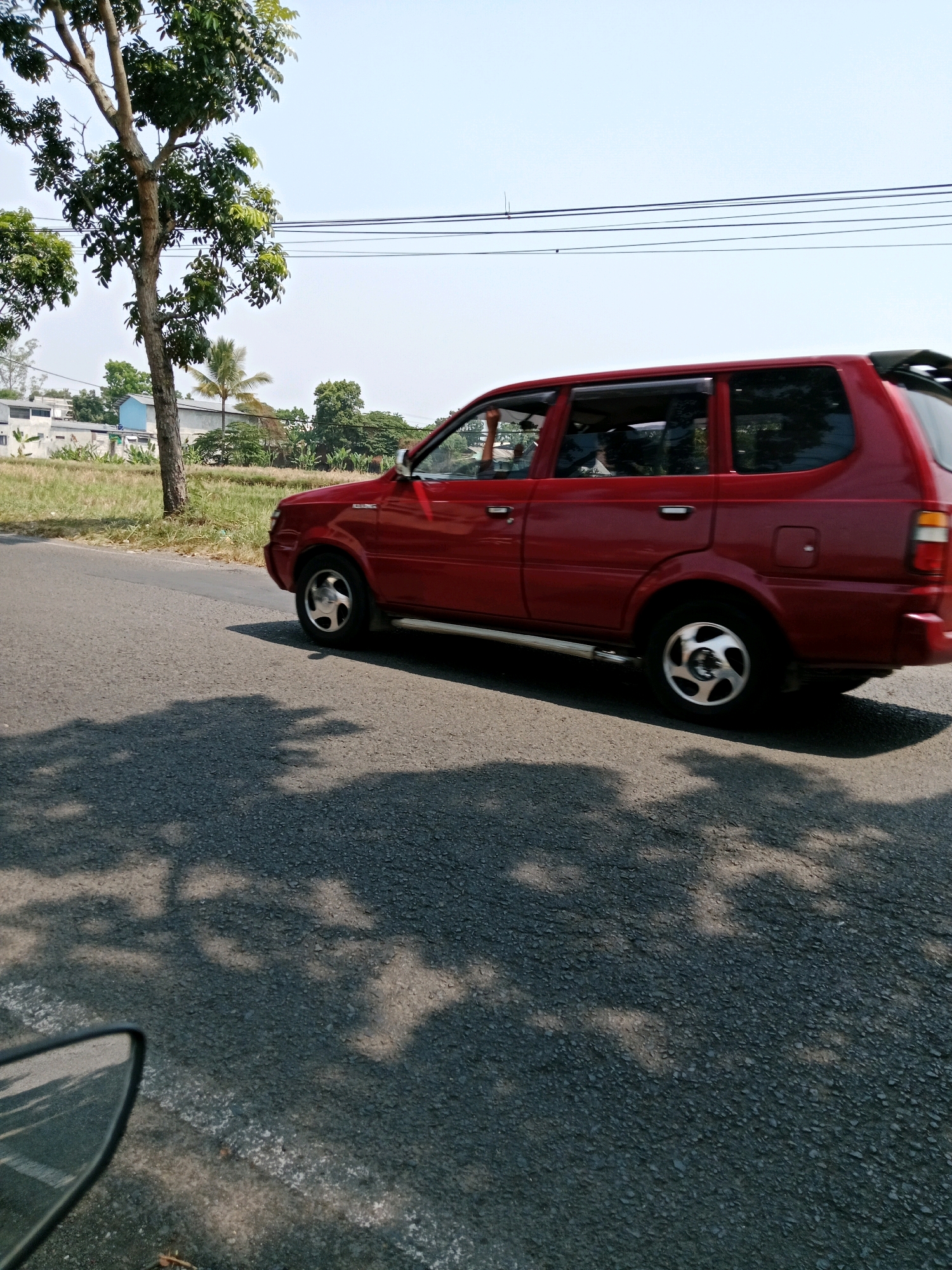 A red SUV is captured in a side profile view, driving on a paved road. The car is in motion, as indicated by the motion blur on the wheels and the road. The environment appears to be a rural or semi-urban area, with vegetation and some buildings visible in the background. The sky is clear and blue, suggesting a sunny day. Shadows of trees are cast on the road, indicating the presence of nearby foliage. The location is identified as Karangtengah, Indonesia.