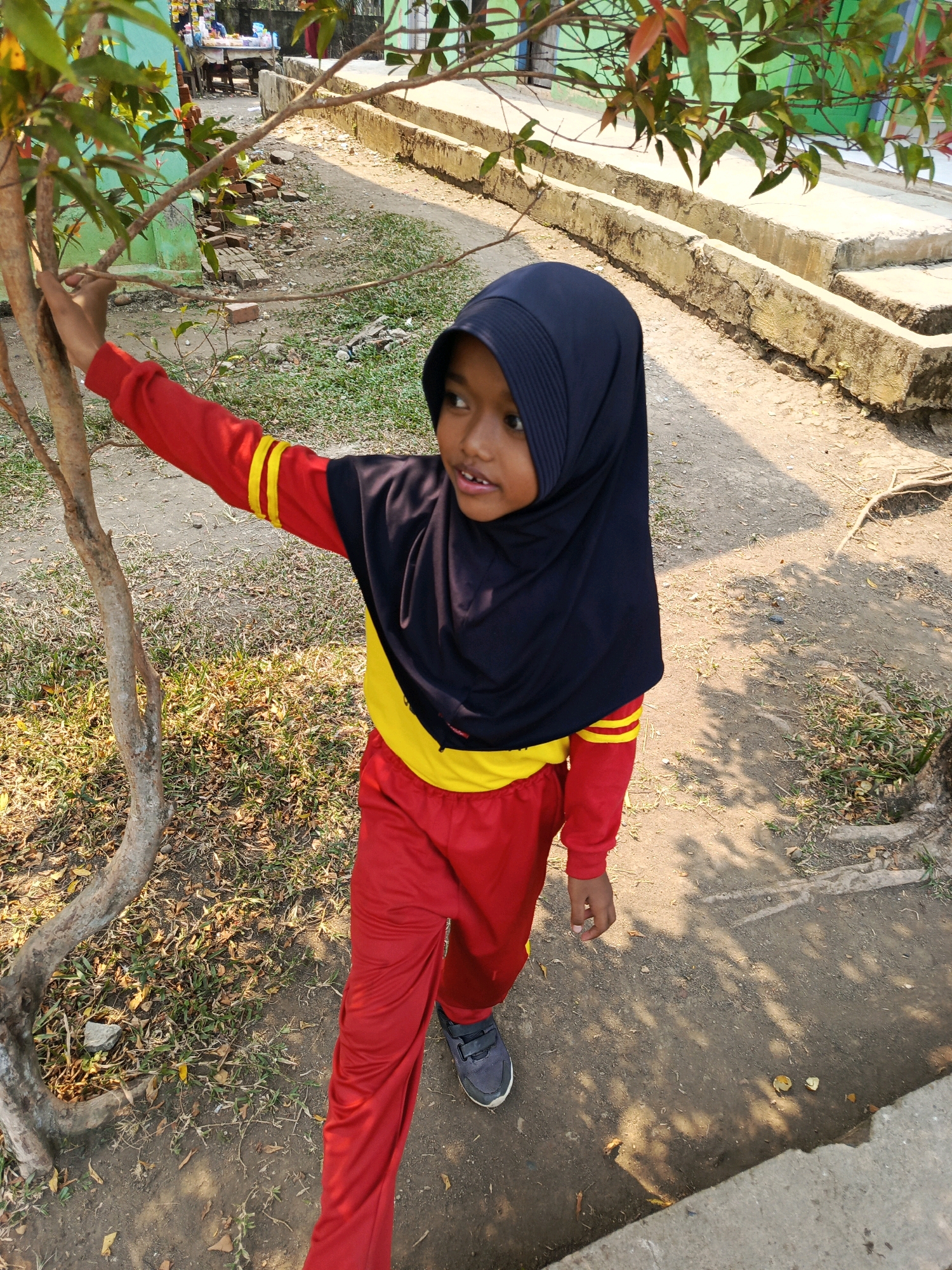 A young girl wearing a dark blue hijab, a red long-sleeved shirt with yellow stripes on the sleeves, and red pants stands outdoors in what appears to be a schoolyard in Megang Sakti IV, Indonesia. She is holding onto the trunk of a small tree with her left hand and looking to her right. The ground is a mixture of dirt and patchy grass. In the background, there are concrete structures, possibly bleachers or a raised walkway, and a building with a green wall. Sunlight casts shadows on the ground, indicating it is daytime.
