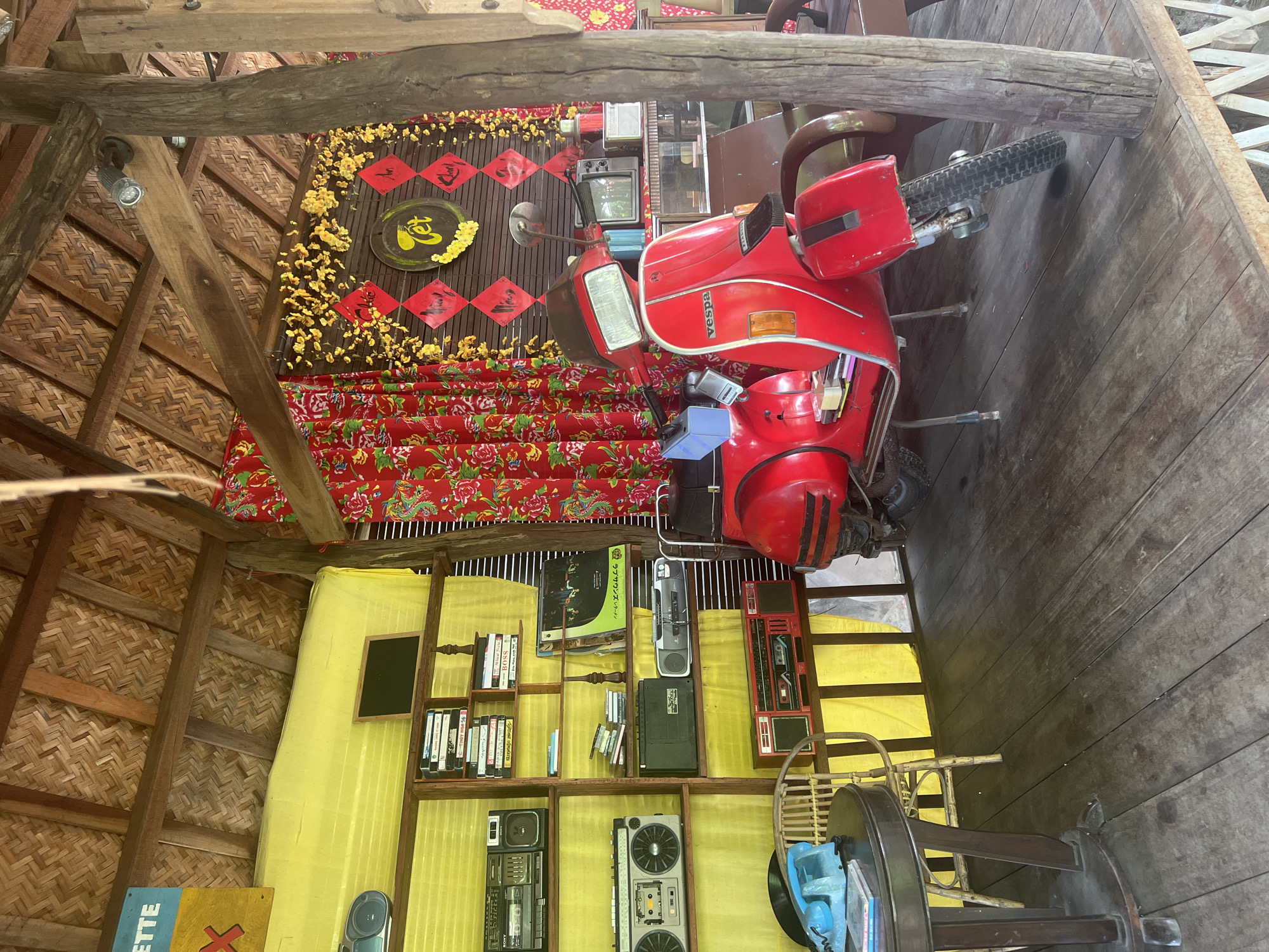 This image depicts a rustic, open-air structure with a thatched roof and wooden beams. The scene is set in Hoi An, Vietnam, as indicated by the Tet (Vietnamese New Year) decorations on the back wall, which include red diamond-shaped banners with Vietnamese calligraphy and yellow floral accents.
Dominating the center of the frame is a bright red Vespa scooter, parked on a wooden floor. To the left of the scooter, a wooden shelf unit displays various vintage electronics, including boomboxes, cassette players, and what appear to be old video tapes or records. A blue retro-style telephone sits on a dark wooden table, accompanied by a wicker chair.
Behind the scooter, there is a glimpse of more wooden furniture and a small television set. The background is adorned with vibrant red curtains featuring a floral pattern. The overall atmosphere is one of nostalgic retro decor, possibly in a shop or a themed cafe. There are no people visible in the image.