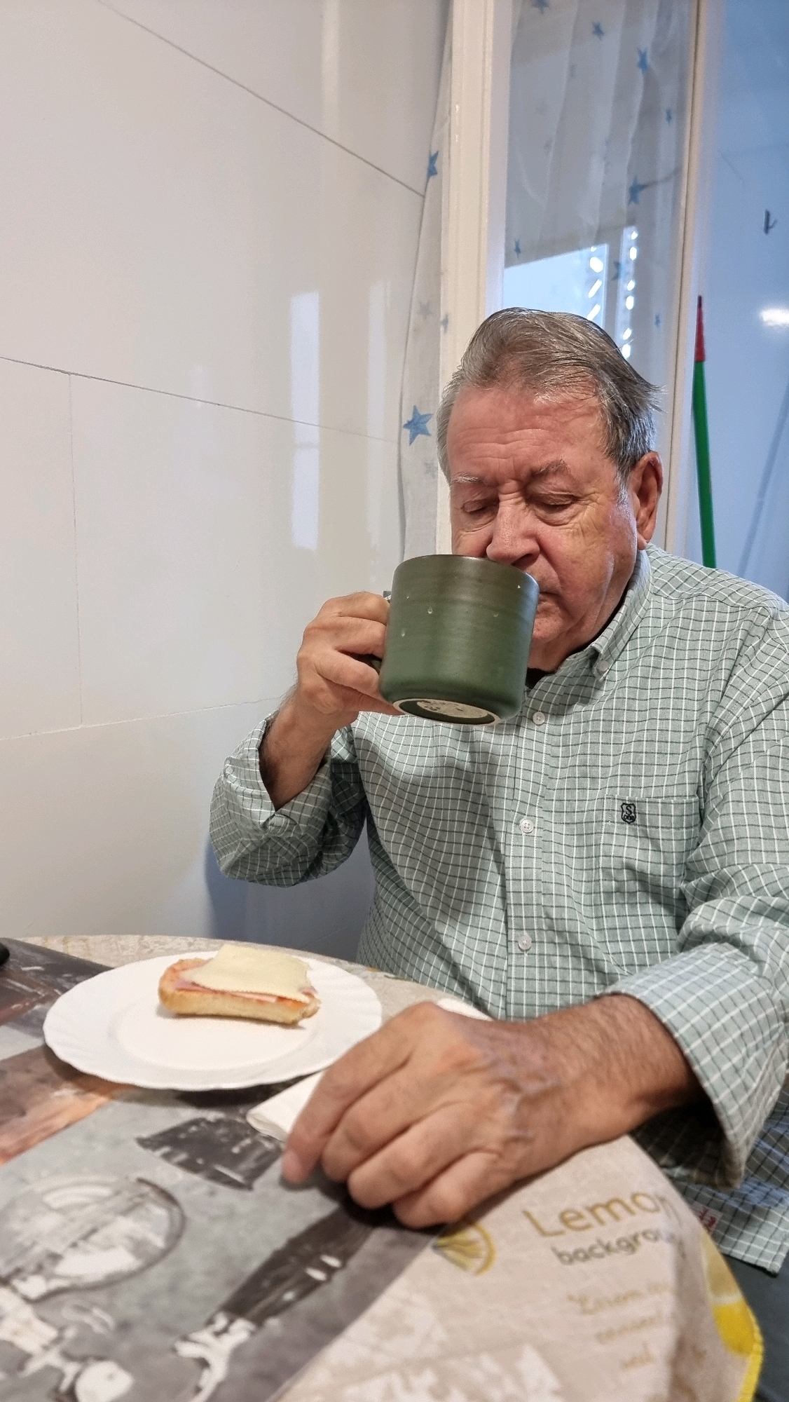 The image depicts an elderly man sitting at a table, holding a green mug and drinking from it. He is wearing a light green checkered shirt. In front of him on a white plate is a small sandwich made with bread, ham, and cheese. The table is covered with a patterned tablecloth. Behind the man, there is a tiled wall and a window with a sheer curtain adorned with blue stars. A green broom with a red tip is visible through the window. The scene appears to be an indoor setting, likely a kitchen or dining area, in Sant Joan Les Fonts, Spain.
