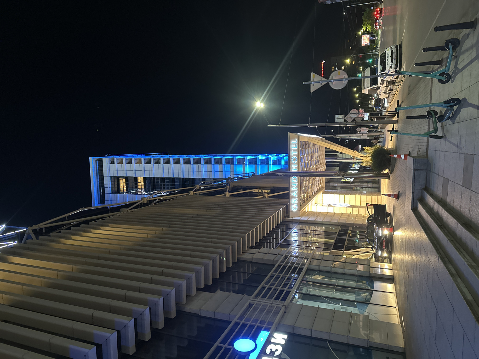 This is a nighttime exterior shot of the Grand Hotel Sofia, located in Sofia, Bulgaria. The hotel's modern facade is illuminated by blue and yellow lights, with a tall, slender tower on the right side of the building glowing with blue light. The entrance area features a covered walkway with a modern design. In front of the hotel, a black Audi sedan with its doors open is parked, with an orange traffic cone nearby. The sidewalk is paved with stone tiles, and a set of concrete stairs leads up to the entrance. To the right of the hotel, a street is visible with traffic, including a white police van and other vehicles. Several electric scooters are parked on the sidewalk in front of the hotel. The sky is dark, indicating it is nighttime.
