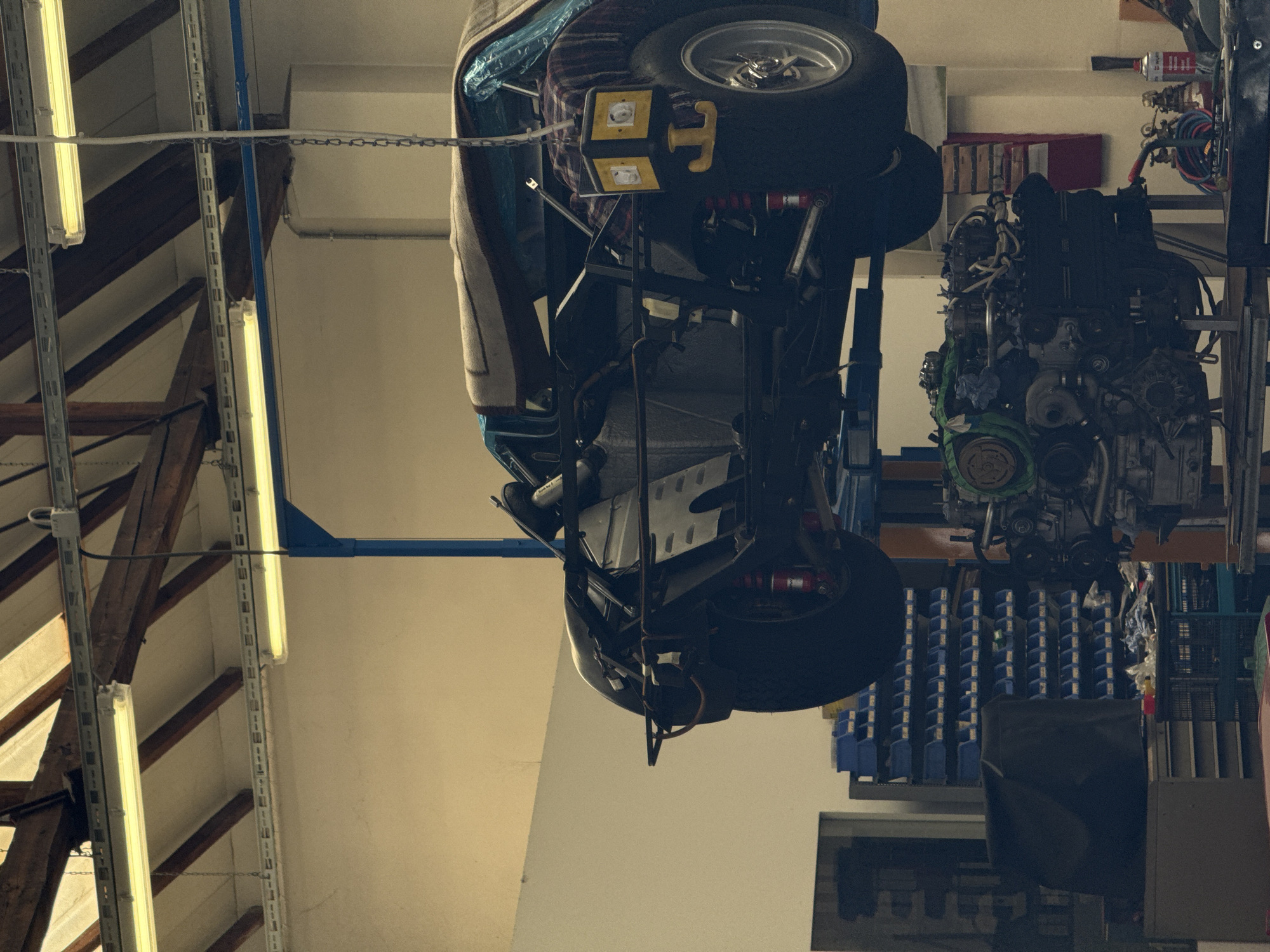 This image depicts a garage scene in Offenbach am Main, Germany, likely a workshop specializing in vehicle repair or restoration. A car chassis, partially covered with blankets and plastic sheeting, is elevated on a hydraulic lift, revealing its intricate internal structure. The car appears to be a sports or racing model, with exposed suspension components and a bare frame. Below the lifted car, a disassembled engine rests on a stand, its various parts, pipes, and wiring clearly visible.
The garage environment is functional and organized, with industrial shelving units filled with blue storage bins lining one wall. Tools and equipment, including a work lamp with yellow accents hanging from a chain, are strategically placed. The lighting comes from fluorescent tubes mounted on the ceiling, casting a bright, even light across the workspace. The background shows a light-colored wall and the wooden beams of the roof structure, typical of an industrial building. The overall impression is one of a professional and dedicated automotive workshop, ready for in-depth mechanical work. There are no people visible in the image.