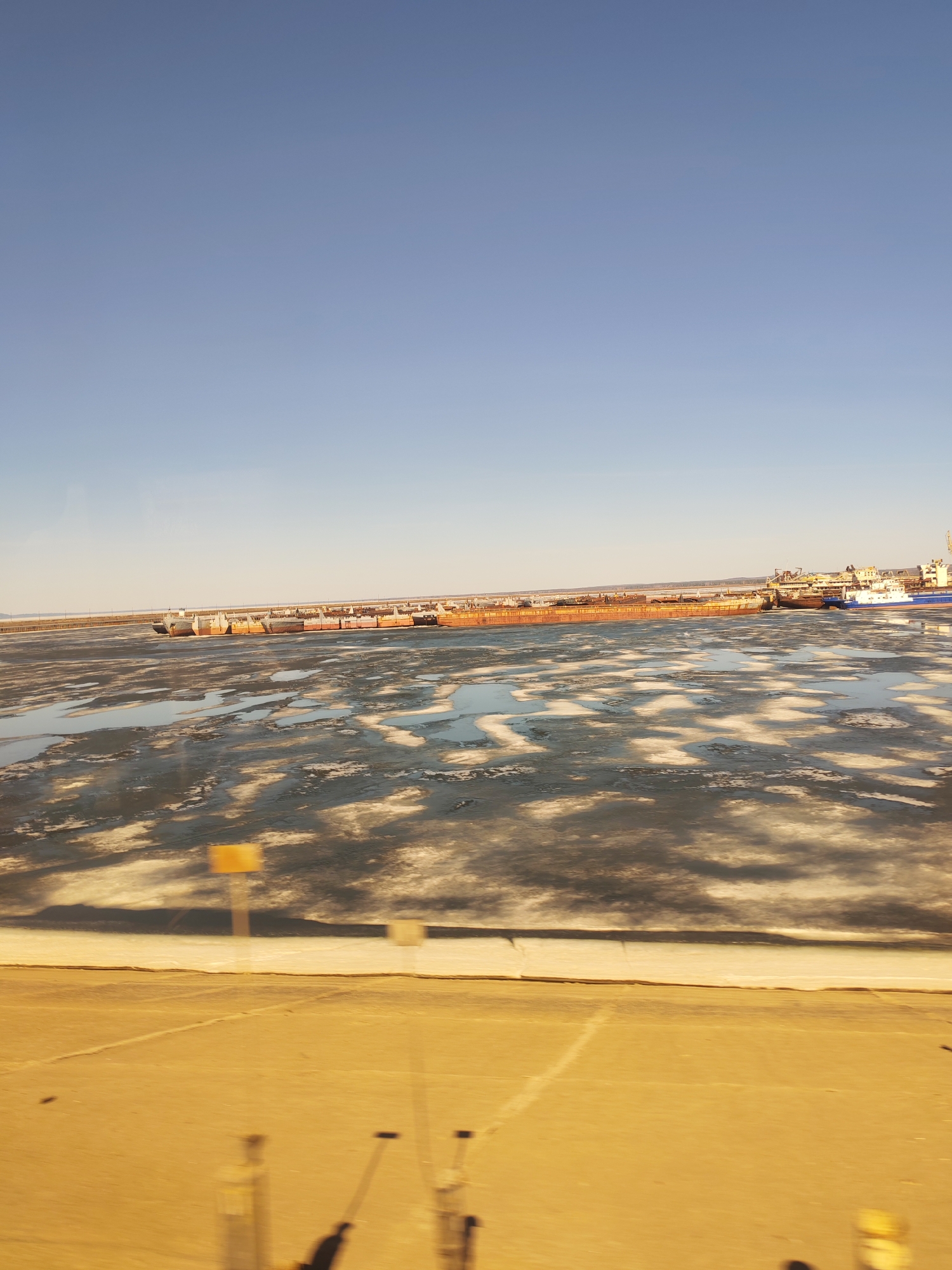 The image shows a view of the Kama River in Chaikovsky, Russia, during a period of partial freezing. The river surface is a mix of ice and open water, with a long line of barges docked along the shoreline in the distance. The sky is clear and blue. In the foreground, there is a paved area that suggests a road or parking lot. The image appears to have been taken from an elevated position, possibly from inside a building or vehicle.