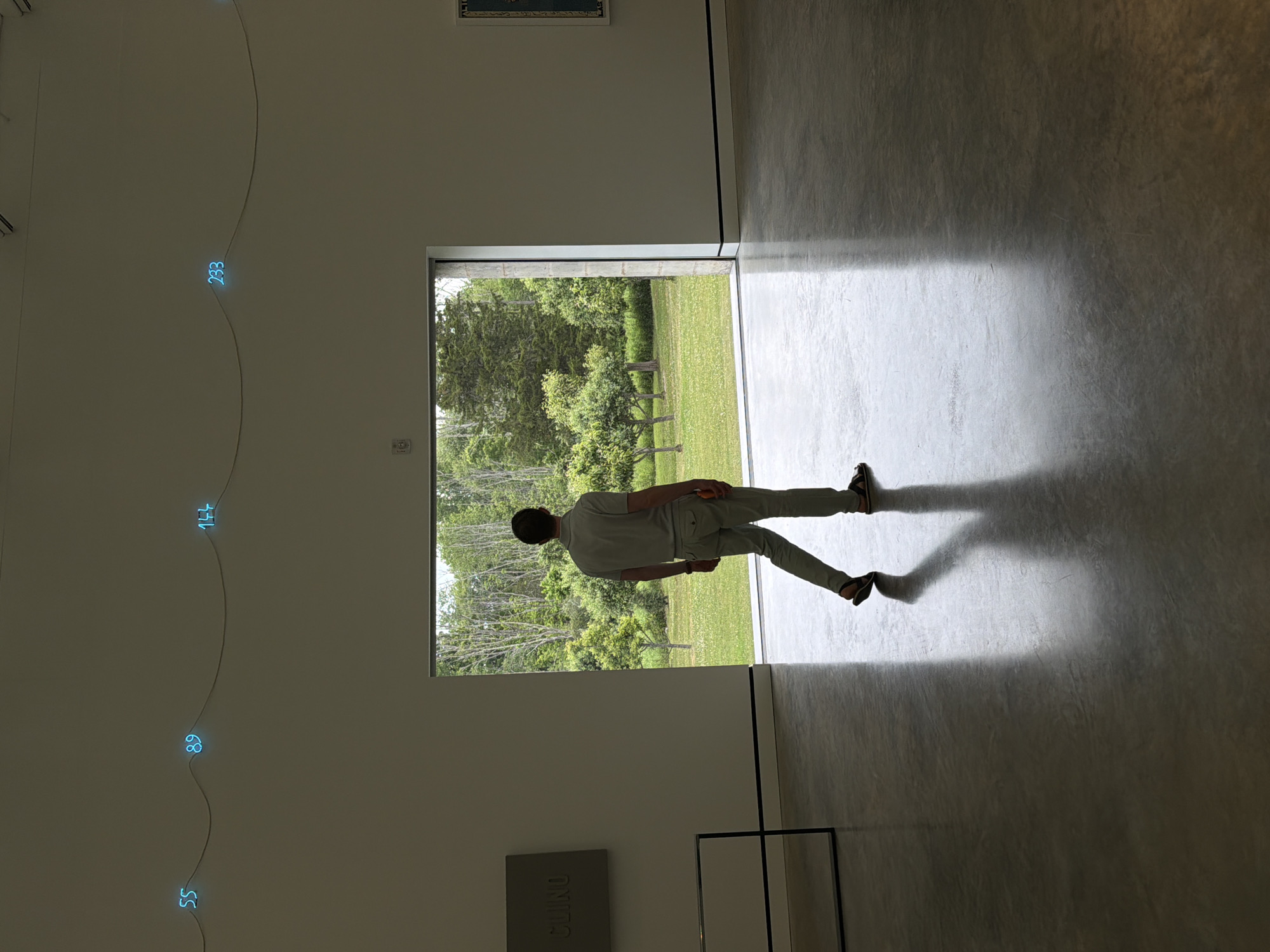 In a contemporary art gallery in Cold Spring, United States, a lone man walks away from the camera towards an open doorway. He is silhouetted against the bright exterior, wearing a light-colored polo shirt and pants, and sandals. The gallery interior features a polished concrete floor reflecting the light. On the wall to the left, a square plaque with the word "CLINO" is visible. Above the doorway, a string of blue neon numbers, "55", "89", "144", and "233", are illuminated against the muted wall. Beyond the doorway, a lush green landscape with trees and a grassy field is visible under a daylight sky.
