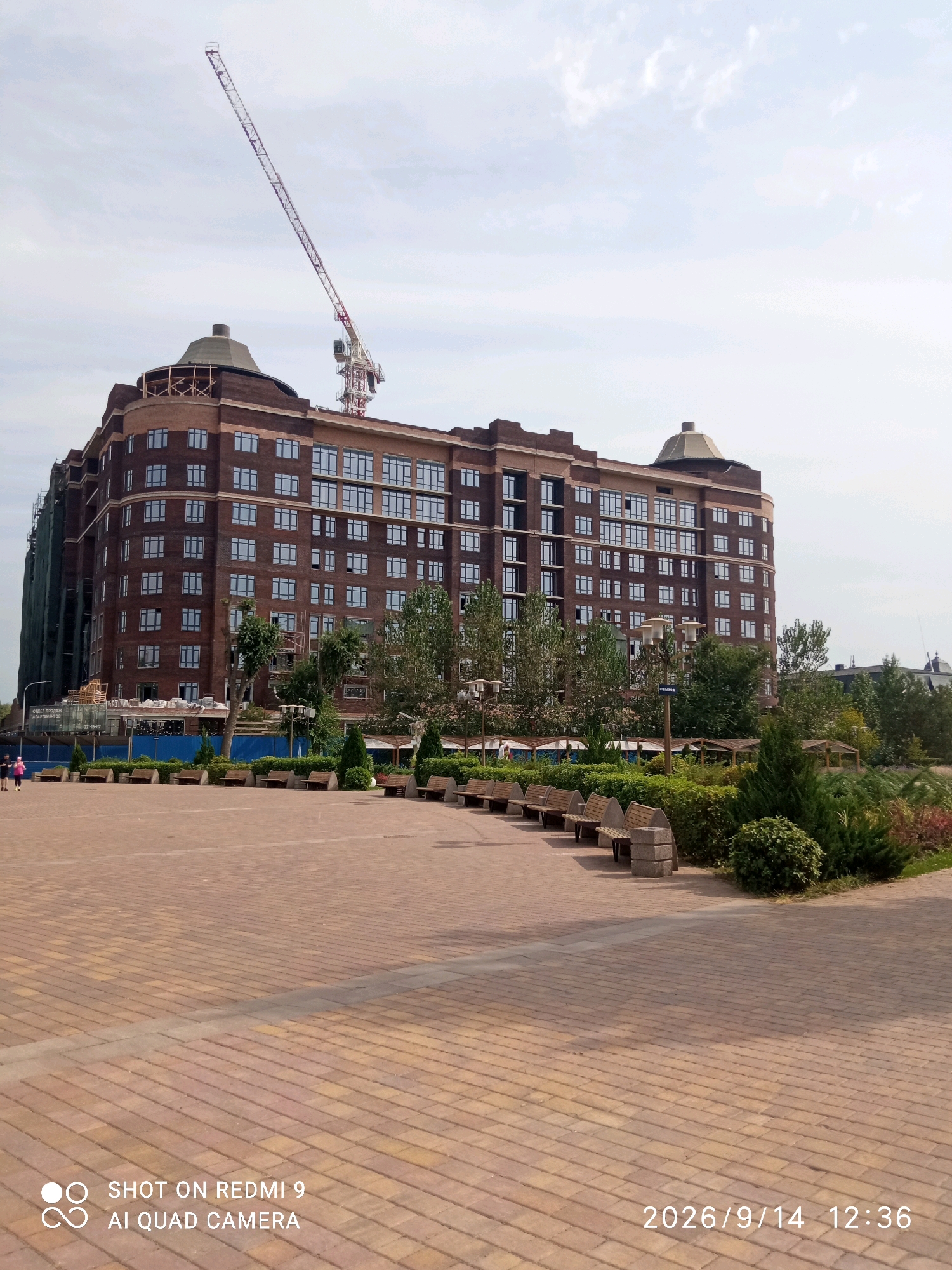 The image captures a wide view of a large, multi-story brick apartment building under construction, located in Rostov-on-Don, Russia. A tall construction crane is visible towering over the building, with its jib extending diagonally across the sky. The building itself features numerous windows and distinctive curved architectural elements at its upper corners, topped with conical structures. Green construction netting is draped over a portion of the building on the left side.
In the foreground, a paved pedestrian area made of interlocking reddish-brown bricks forms a plaza. Several benches are arranged along the edge of this plaza, facing a landscaped area with trees, bushes, and some small covered gazebos. A streetlamp is also visible in the landscaped area. Two individuals, a woman in a pink shirt and black shorts and another person in a black outfit, are walking away from the camera on the left side of the plaza. The sky is overcast with scattered clouds. A timestamp and "SHOT ON REDMI 9 AI QUAD CAMERA" watermark are present in the lower left and right corners of the image, respectively. The date displayed is 2026/9/14 12:36.