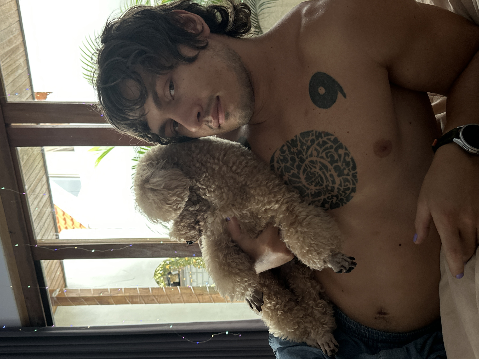 A shirtless man with dark, wavy hair is holding a brown, curly-haired dog against his chest. He is looking at the camera with a slight smile. He has tattoos on his chest, including one that resembles the number 6 and a circular design. He is wearing a watch on his left wrist and blue jeans. A hand with lavender painted nails is visible near the dog. The background includes a window with string lights, glimpses of greenery, and a wooden structure. The location is not clear from the image, therefore the location cannot be confirmed to be in Dalung, Indonesia.
