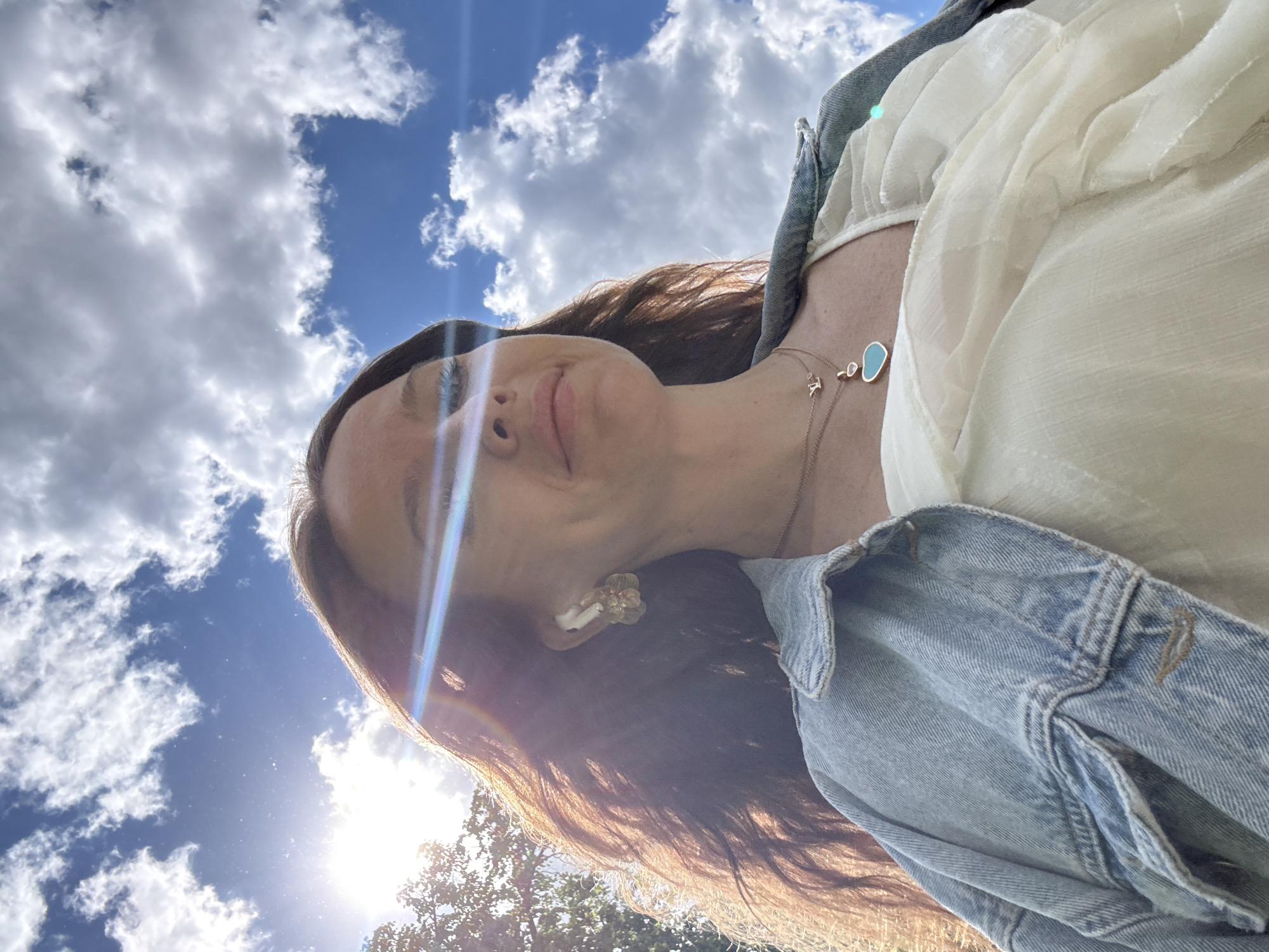 The image captures a low-angle perspective of a young woman looking upwards towards a bright, cloud-filled sky. She has long, dark brown hair and fair skin, with a serene or slightly smiling expression on her face. Her right eye is particularly visible, and a bright lens flare from the sun passes over her features.
She is dressed in a light-colored, possibly white or cream, top with a textured, flowy fabric, worn under a light blue denim jacket. Her accessories include a delicate gold necklace featuring two charms: one is an oval-shaped, light blue or turquoise pendant, and the other is a smaller, thinner gold chain with a minuscule "LV" initial charm. She also wears a decorative earring, possibly floral, and a white earbud in her right ear.
The background consists of a vibrant blue sky dotted with scattered, fluffy white clouds. The sun, positioned in the lower right (when viewing the image in its intended upright orientation), is intensely bright, causing a prominent lens flare across the image, especially over the woman's hair and face. The tops of some trees, mostly silhouetted by the sun, are visible in the lower right corner, suggesting an outdoor setting like a park or garden. The overall impression is one of bright daylight and pleasant weather. Given the provided context, the scene is located in Moscow, Russia.