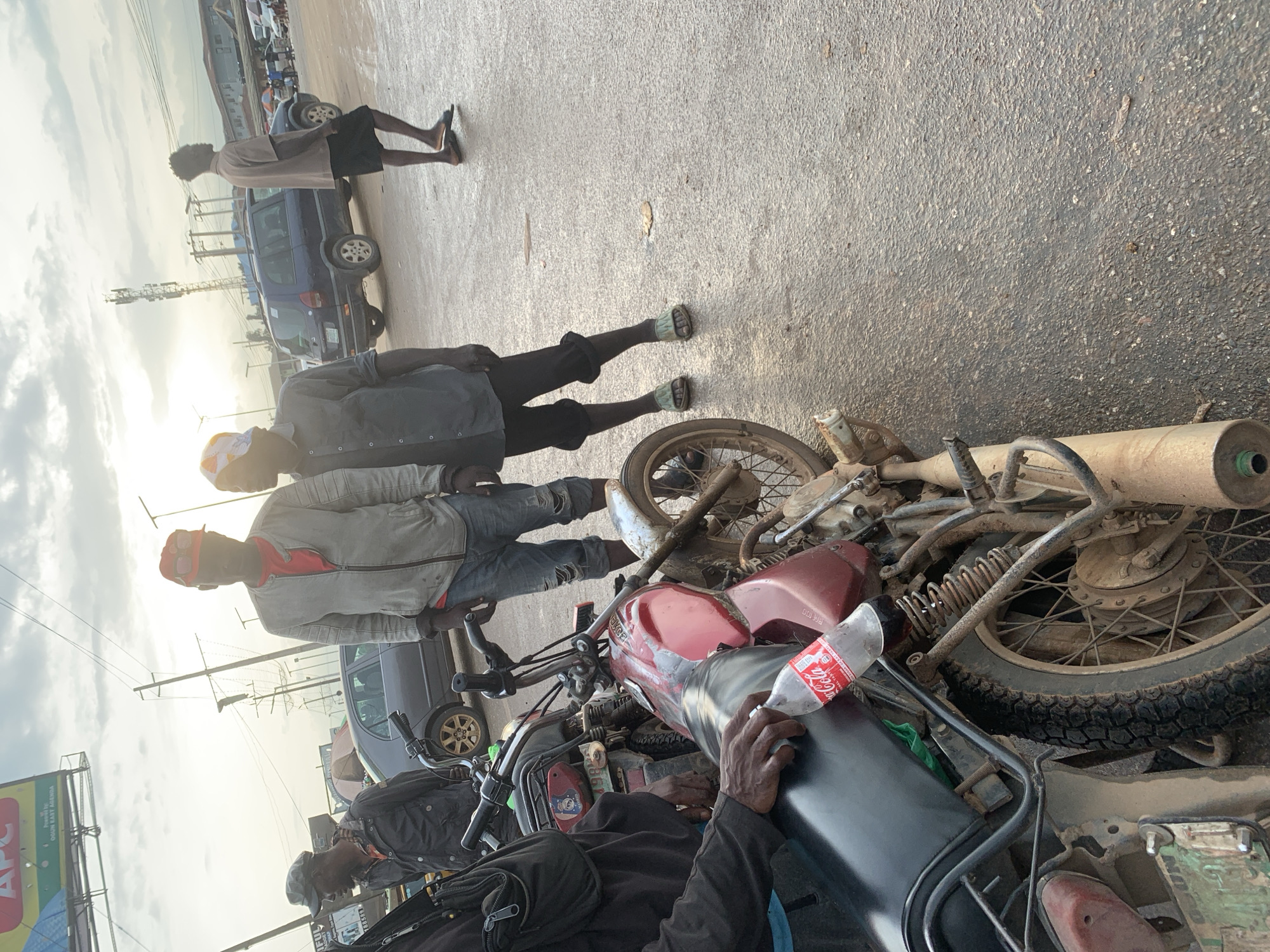 In Shagamu, Nigeria, a group of people are gathered near parked motorcycles and vehicles under an overcast sky. A red Coca-Cola bottle is precariously balanced on the seat of a red motorcycle in the foreground, its cap open. The motorcycle is weathered and dusty, with a visible license plate. Two men stand next to the motorcycle, one wearing a red cap and light-colored jacket, the other in a dark shirt and shorts. Further back, other individuals and vehicles, including a dark blue SUV and a grey sedan, are visible. The ground is paved with asphalt, showing signs of wear and tear. The overall scene suggests a typical street setting in the city.