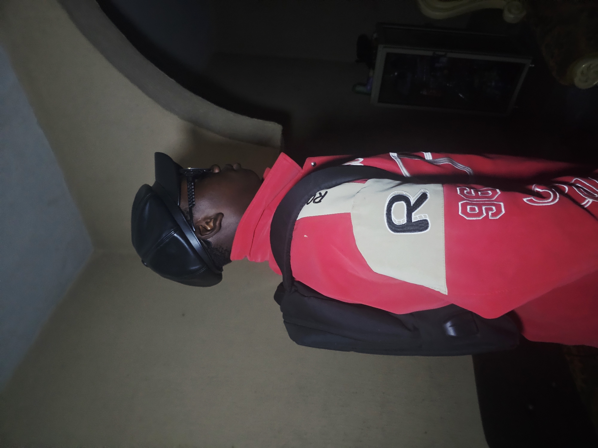 An individual wearing a black cap, sunglasses, a red jacket with white and black accents, and carrying a black bag is observed in an indoor setting. The scene appears to be in Nkwelle, Nigeria. The environment is dimly lit. The subject's pose is mostly sideways to the camera. A wall and a ceiling fixture are also visible.