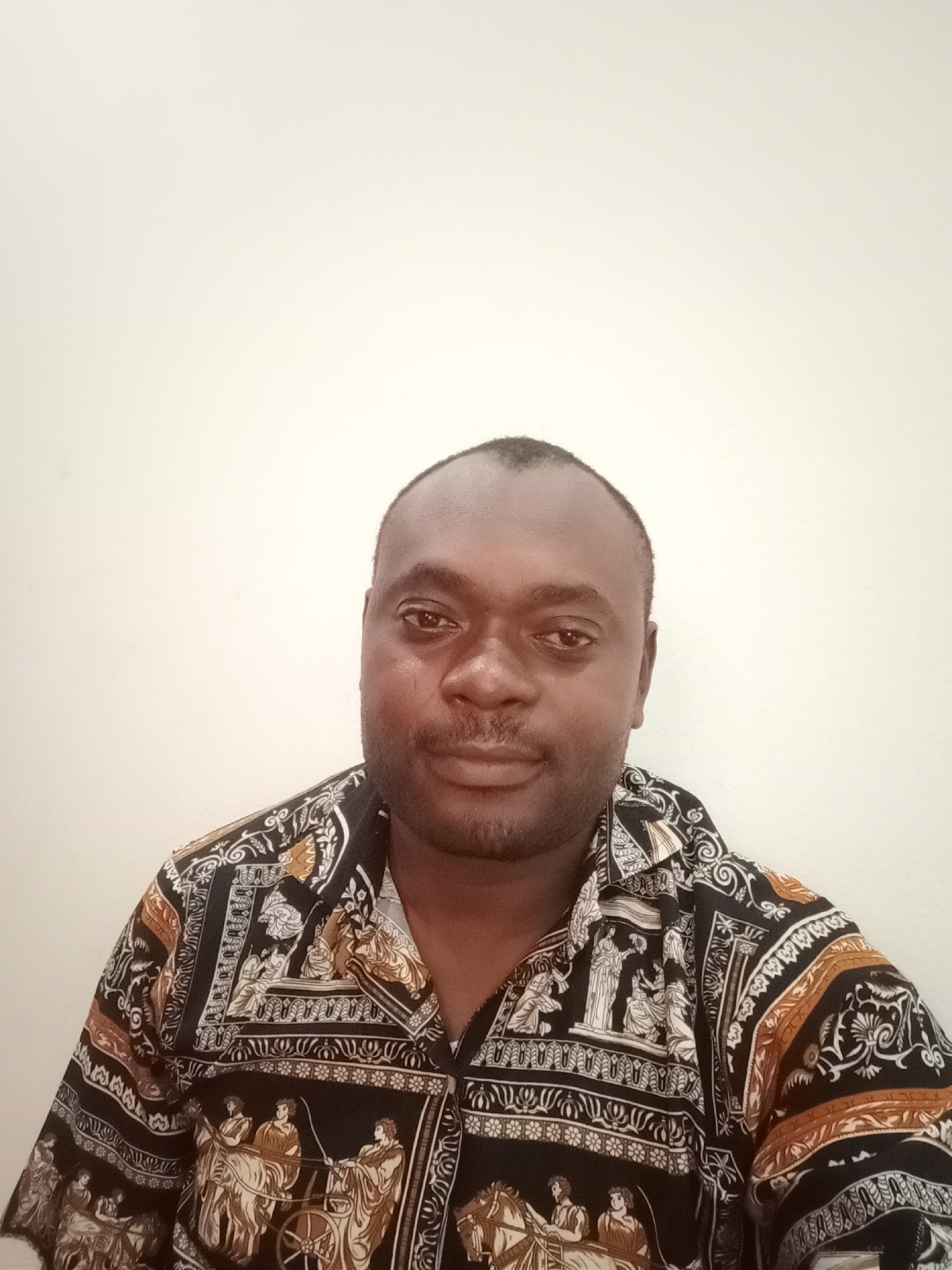 A close-up studio-style shot shows a man centered against a plain, off-white wall. He has dark skin, short black hair, and a neatly trimmed beard and mustache. The man is wearing a patterned shirt with a black background and images of Roman or Greek figures and chariots in white, gold, and tan. The style of the shirt suggests it is a vintage or retro design. There are no clear indicators of the location being Gwarinpa, Nigeria, in the immediate visual context.