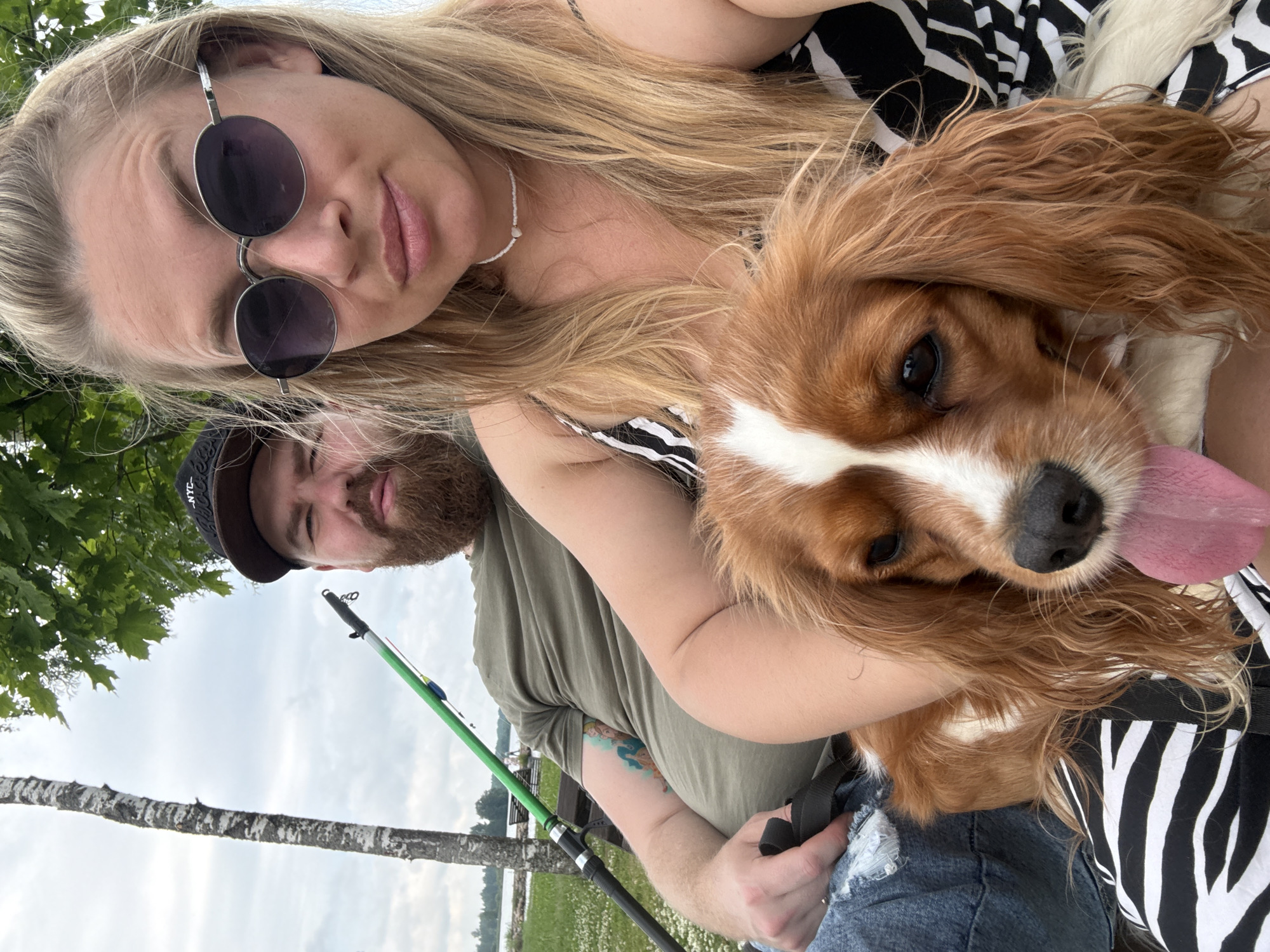 A close-up selfie shows a woman holding a Cavalier King Charles Spaniel dog. The woman has blonde hair and is wearing sunglasses and a black and white striped top. The dog is light brown and white with its tongue hanging out. In the background, a man with a beard and a baseball cap is visible, holding a fishing rod. They appear to be sitting on a bench by a lake, with a birch tree and green foliage surrounding them. The scene is set outdoors, likely in a park or recreational area in Valday, Russia, indicated by the presence of fishing equipment and the natural landscape. The overall mood is casual and relaxed.
