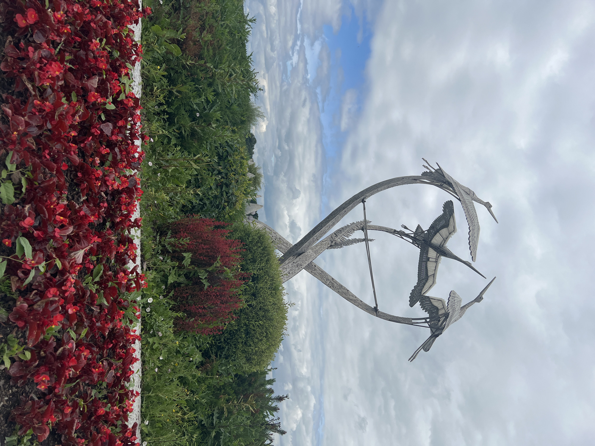 The image shows a sculpture of four cranes in flight, made of weathered metal. The sculpture is situated in a garden with lush green foliage and a bed of bright red flowers in the foreground. The sky is overcast with scattered clouds. The sculpture is located in a park in Minsk, Belarus, and is a popular spot for photography.