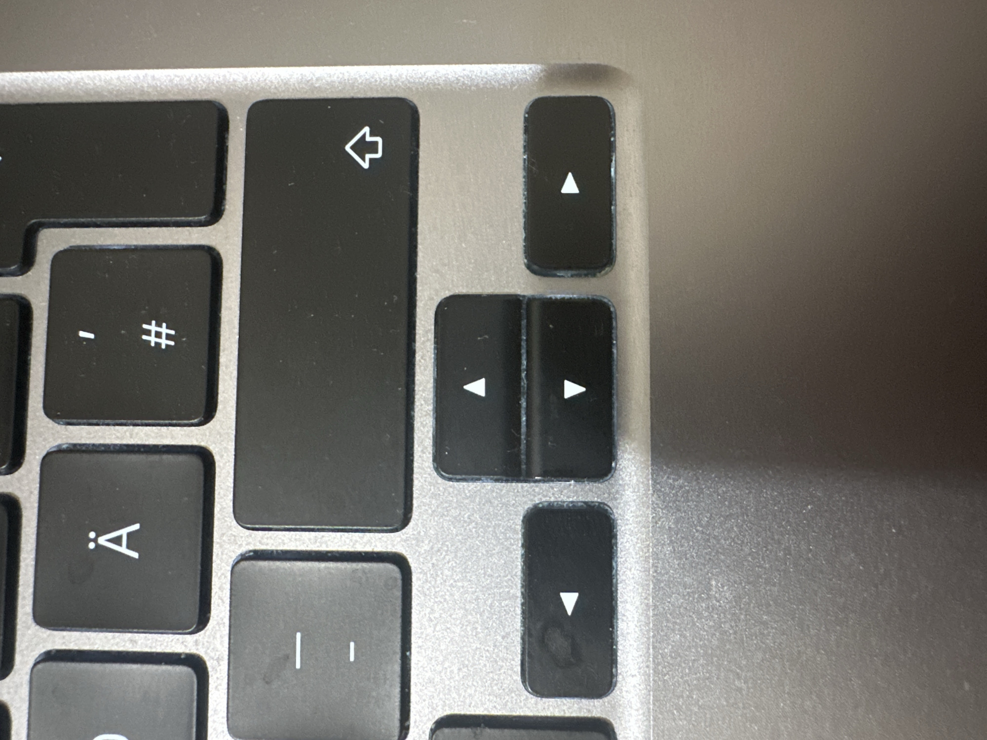 The image shows a close-up view of a portion of a laptop keyboard. The keyboard is dark gray with white lettering. Visible keys include those with the characters "Ä", "#", "-", and arrow symbols pointing left, up, down, and right. The keys show signs of wear, with some scuffing and faint smudges on their surfaces. The surrounding area of the laptop is metallic gray and appears to be made of aluminum. The lighting suggests it is an indoor setting, and there is no indication of the laptop's location in Hamburg, Germany, or any people, actions, or interactions.