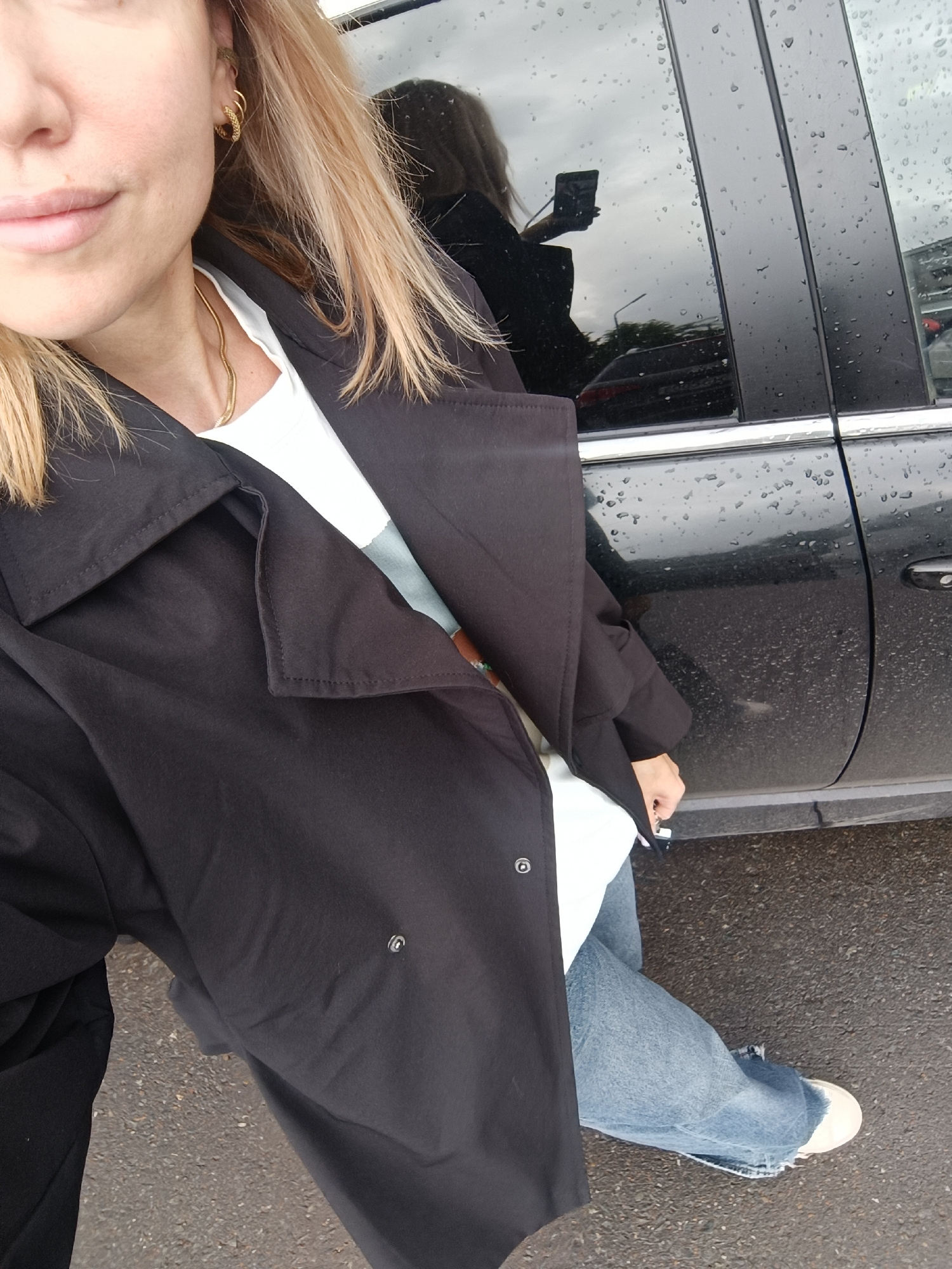 A high-angle selfie captures a person standing outdoors next to a dark-colored car, likely in Naberezhnye Chelny, Russia. The subject, a woman with light blonde hair, is partially visible from the chest up, showing her fair skin, neutral-toned lips, and a portion of her face. She accessorizes with several gold hoop earrings, one appearing braided or textured, and a thin gold snake-chain necklace.
Her outfit includes a black, wide-collared trench coat or jacket worn over a white t-shirt. She is also wearing light-wash, wide-leg or flared denim jeans with a distressed hem and light-colored sneakers with white soles.
The activity taking place is a selfie, as evidenced by the clear reflection in the car's rear window, showing the woman holding a smartphone to capture the image. The scene is set outdoors on what appears to be asphalt ground. Notable details include the weather conditions: the car's windows and body are covered in numerous raindrops, indicating rain or recent wet weather. The reflection in the car window also shows an overcast sky and faint outlines of distant structures or trees. The time of day appears to be daytime, under cloudy conditions. There is no visible text in the image.