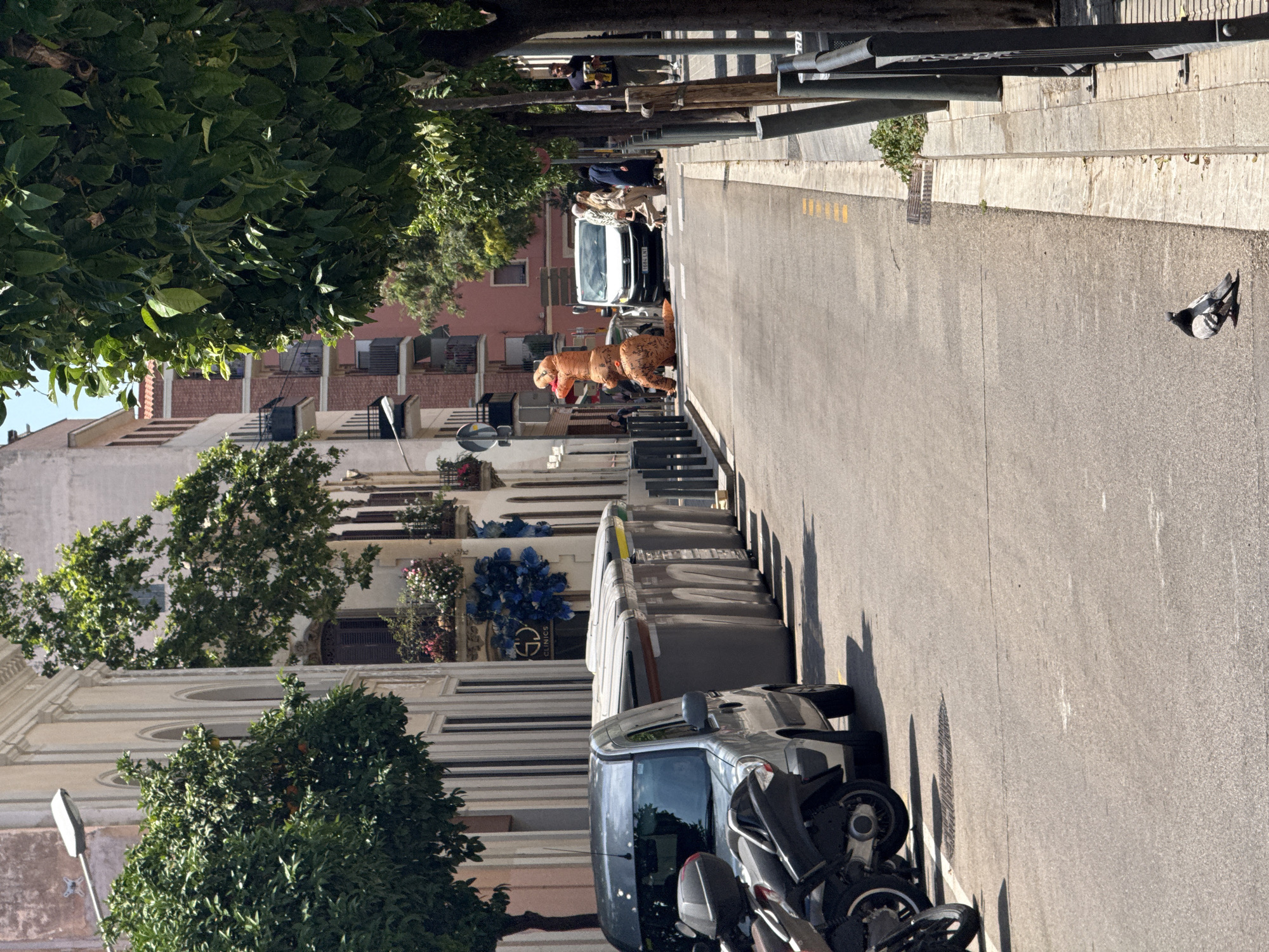 The image depicts a street scene in L'Hospitalet de Llobregat, Spain. The perspective is from street level, looking down a two-lane road lined with buildings and trees. In the foreground on the left, a silver van and two motorcycles are parked. A row of large, grey trash receptacles runs along the sidewalk. Further down the street, a person in an inflatable dinosaur costume is walking across the road, in front of a white van. Several other pedestrians are visible in the middle ground, including a man walking a dog. The buildings appear to be a mix of residential and commercial, with balconies and architectural details. Trees with green foliage line both sides of the street. The sky is clear and blue, indicating a sunny day. A pigeon is seen on the asphalt in the lower right of the frame.