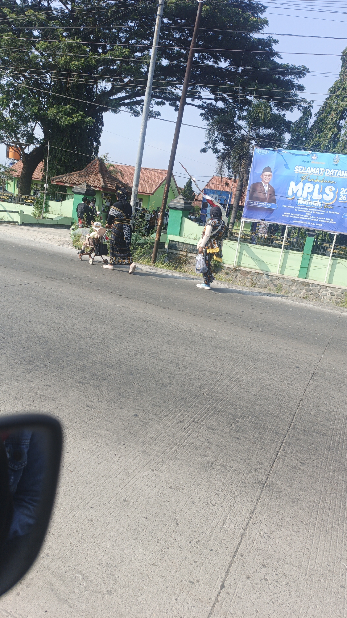 The image depicts a street scene in Sumengko, Indonesia, under a clear, bright sky. A group of people are walking along the side of a paved road. Two individuals in traditional attire, one with a black and gold patterned garment and the other with a similar outfit adorned with yellow and white accents, are prominent. The person in the black and gold outfit appears to be pushing a small stroller. Another individual is walking behind them carrying a plastic bag. In the background, there are green buildings with orange-tiled roofs, a large tree with dense foliage, and a blue banner displaying text and an image of a man in formal attire. The banner appears to announce an event. A utility pole stands near the center of the image. The road has visible tire tracks and expansion joints. The reflection of a person wearing blue jeans and a dark jacket is visible in the lower-left corner of the image, suggesting the photo was taken from a vehicle.