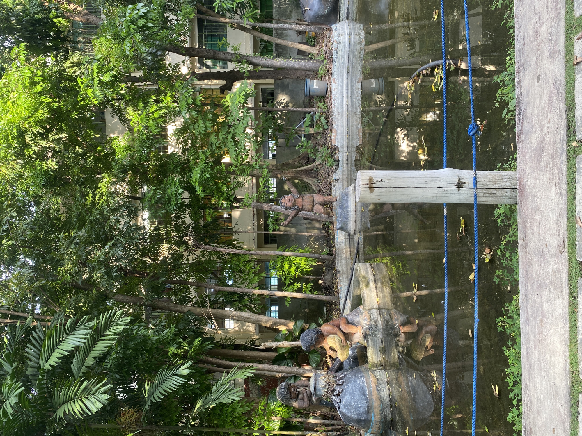 The image shows a pond with several statues of children in and around it. The pond is surrounded by lush greenery, including palm trees and other foliage. In the background, a building with windows can be seen. The pond is bordered by a concrete edge and a blue rope fence. The water reflects the surrounding trees and the building. The statues depict children engaged in various activities, such as playing with a basket and one appearing to be pouring water. The overall scene suggests a park or garden area. This location is identified as the Peoples' Park in Davao City, Philippines.