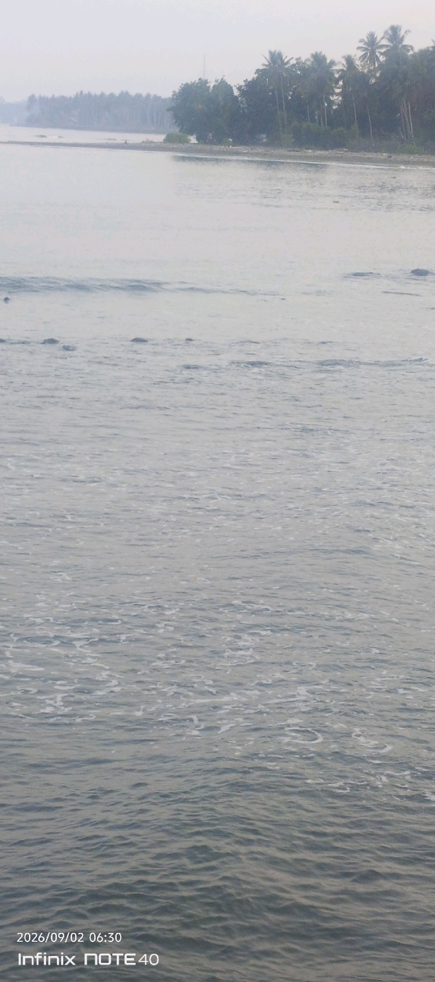 A calm ocean scene in Malunda, Indonesia, with a light haze obscuring the distant treeline. The water is a muted grey-blue with gentle ripples and some scattered white foam. In the foreground, the water is slightly choppier with more visible foam patterns. Along the distant shore, a dense line of trees, including palm trees, forms a dark silhouette against the hazy sky. The overall impression is one of a serene, overcast day at the coast. The lower right corner of the image displays a timestamp: "2026/09/02 06:30" and device information: "Infinix NOTE 40".